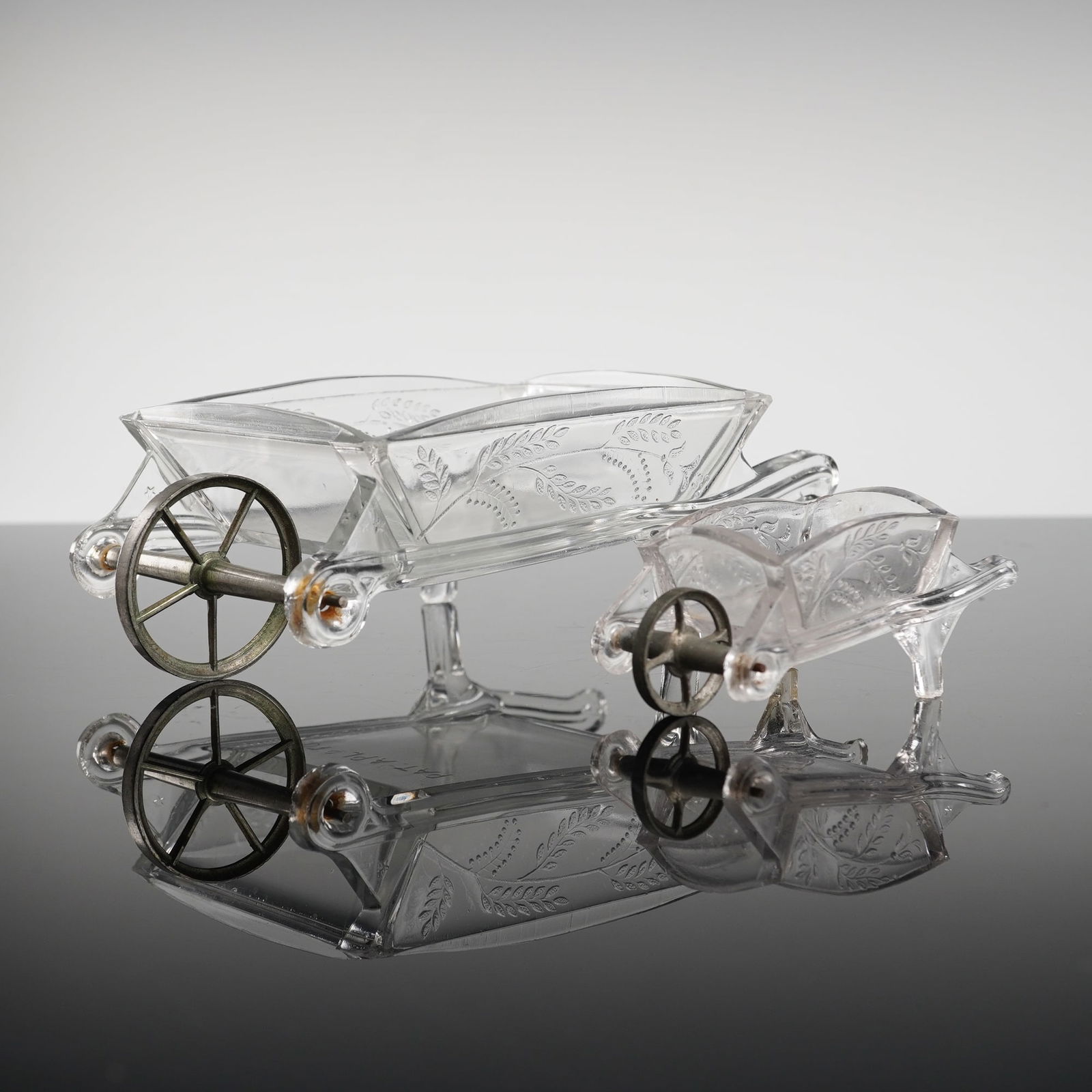 A Pair of EAPG Wheelbarrow Novelty Dishes: Pressed glass novelty dishes, consisting of one large and one small wheelbarrow. These wheelbarrows feature clear molded glass bodies decorated with a repeating floral and vine pattern along the sides