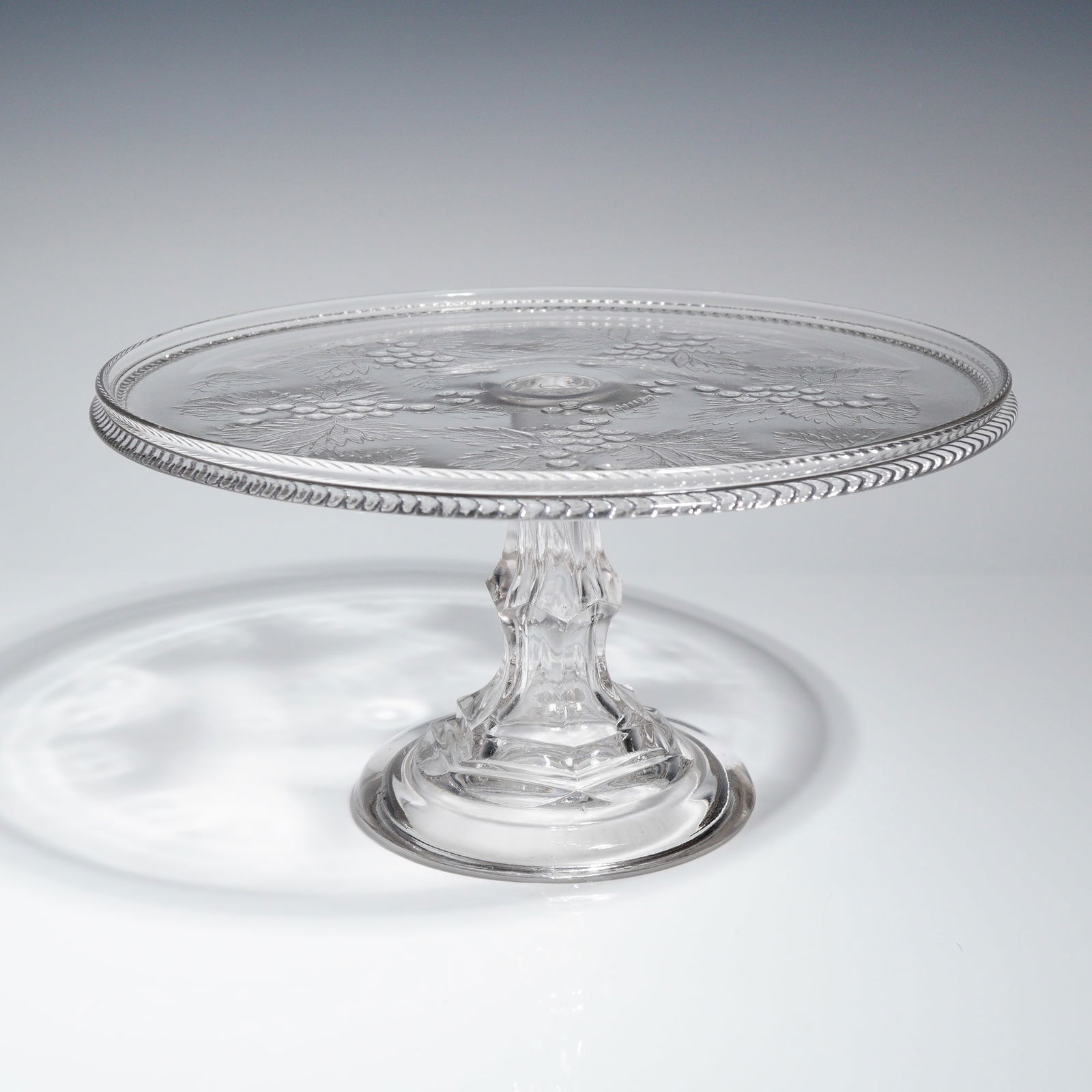 Adams & Co. EAPG Good Luck Pattern Pressed Glass Pedestal Cake Stand: A single Early American Pressed Glass (EAPG) pedestal cake stand manufactured by Adams & Co. in the 'Good Luck' pattern, also known as 'Leaf & Berry'. The object is constructed from clear, colorless p