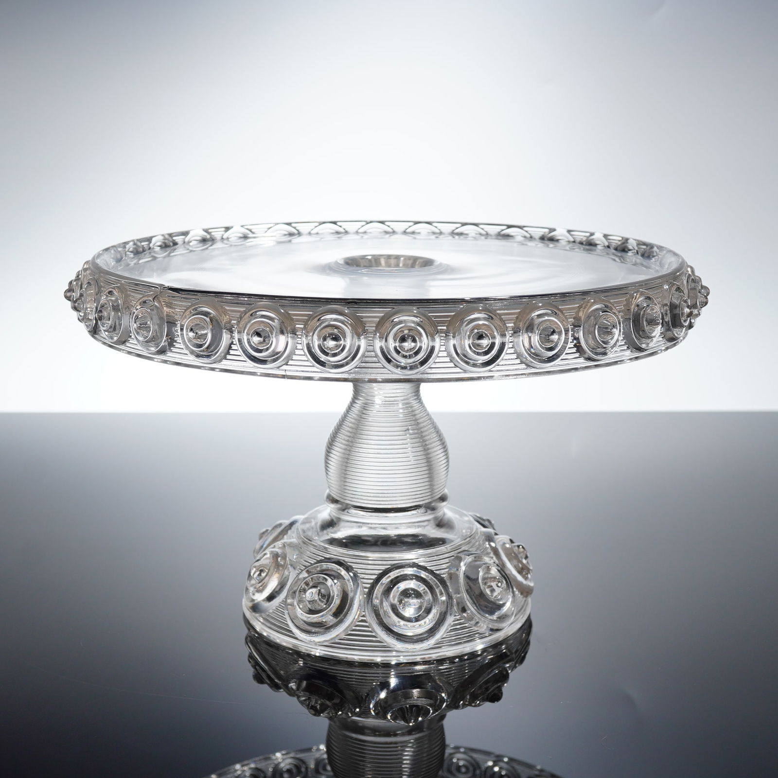 LE Smith Moon and Stars Clear Pressed Glass Cake Stand: A single Early American Pressed Glass (EAPG) cake stand in the "Moon and Stars" pattern, commonly attributed to L.E. Smith Glass. This clear pressed glass pedestal dish features a shallow circular pla