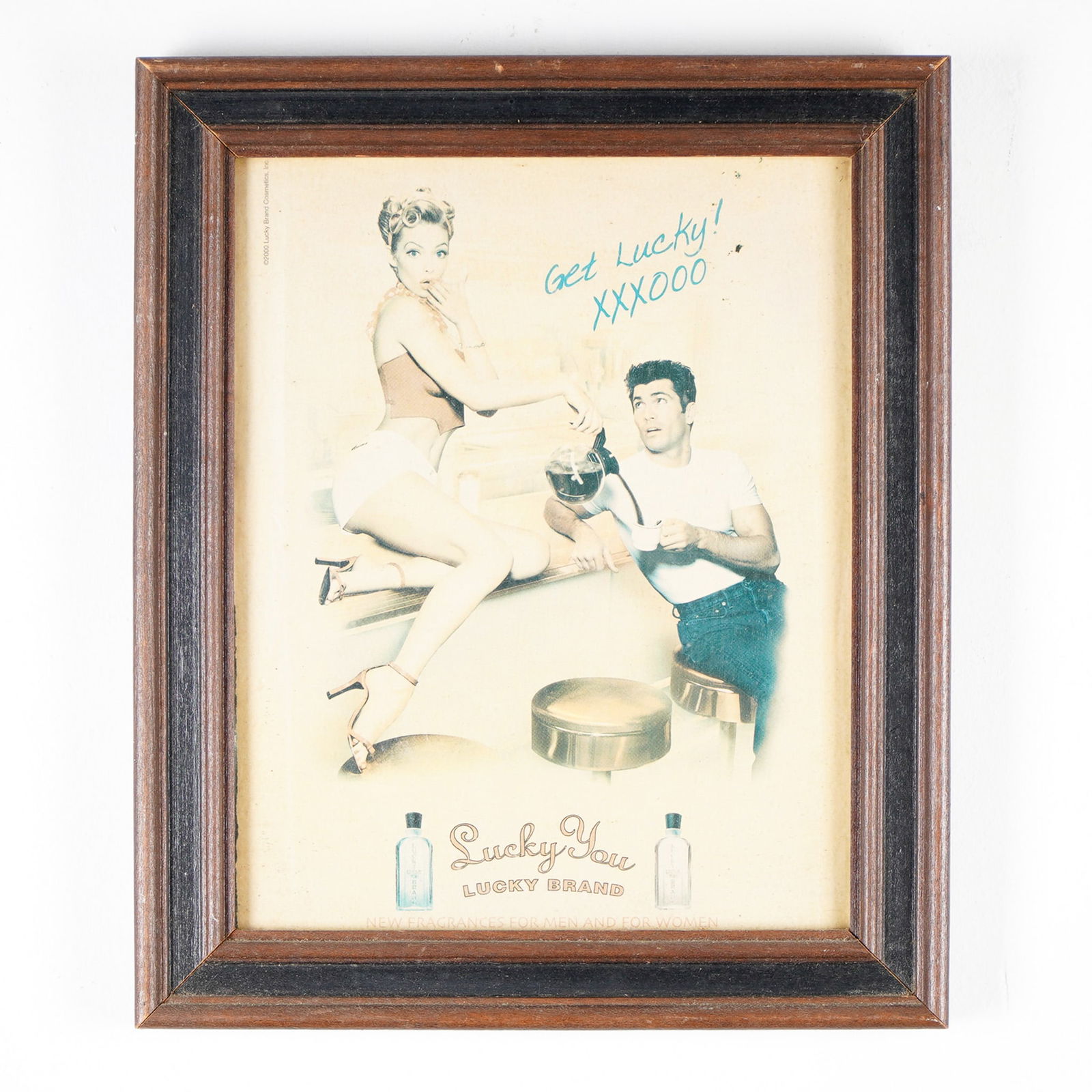 Lucky Brand "Lucky You" Framed Ad Poster: Framed vintage-style advertisement poster for Lucky Brand's "Lucky You" fragrance, dated 2000. The artwork features a retro-inspired diner scene with a pin-up style waitress pouring coffee for a man,