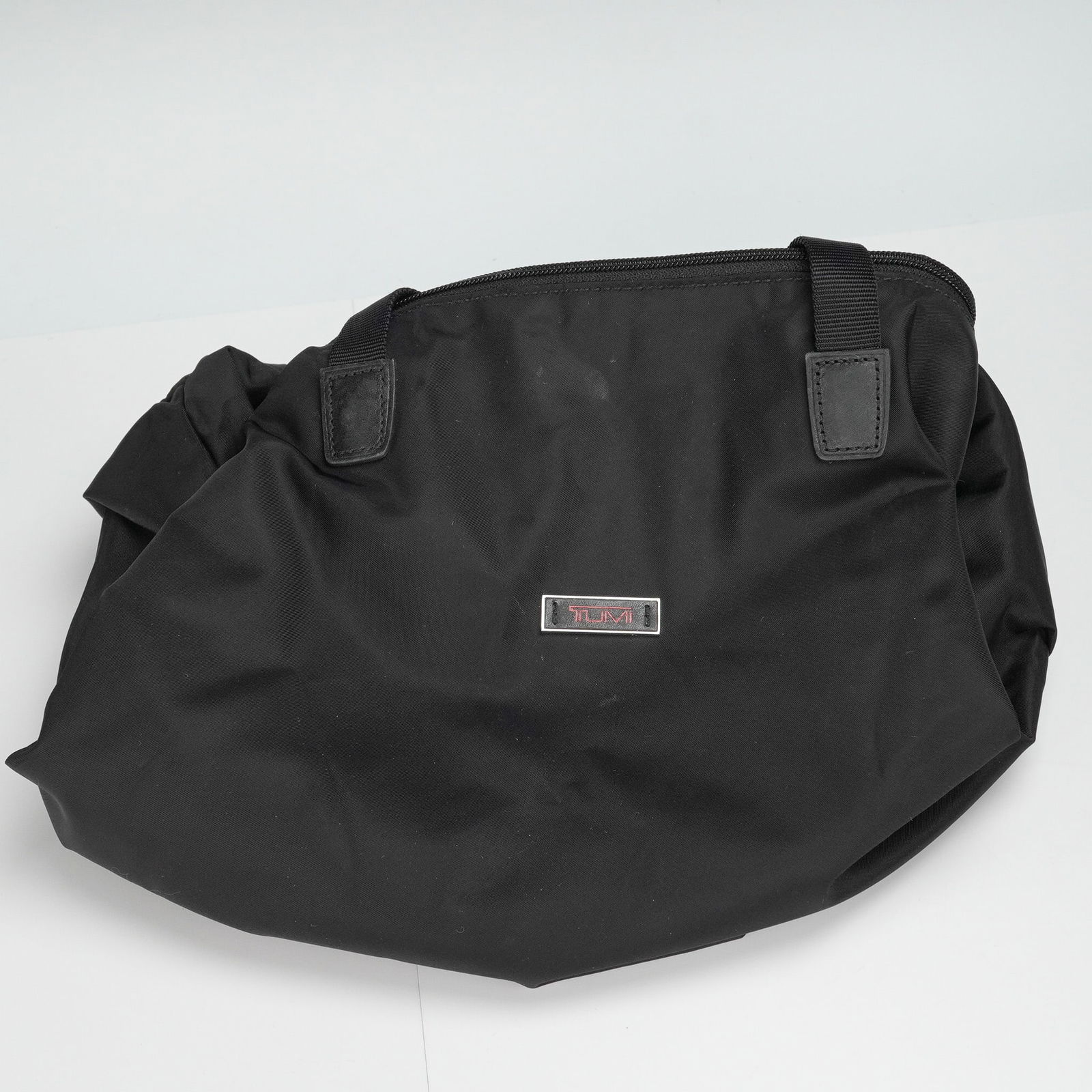 Tumi Black Nylon Travel Tote: A black travel tote featuring dual top handles, a top zip closure, and a spacious interior. The exterior includes a stitched slip pocket and a Tumi logo plaque in polished metal. The tote is crafted f