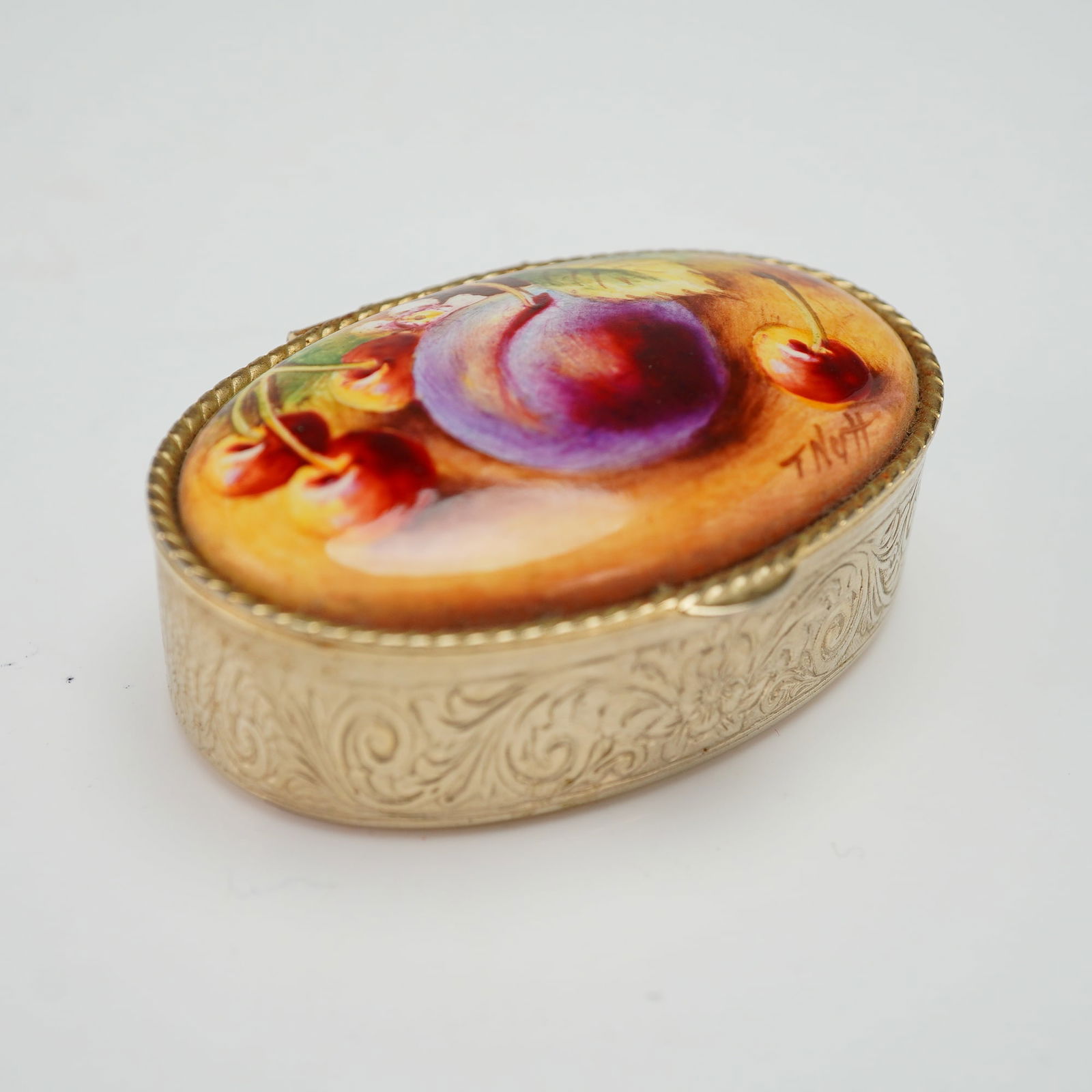 T. Nott Floral and Fruit Enamel Curio Box: An oval metal box designed by artist T. Nott featuring a hinged lid decorated with a hand-painted still life composition of a plum and cherries. The box surfaces exhibit cast floral patterns and folia