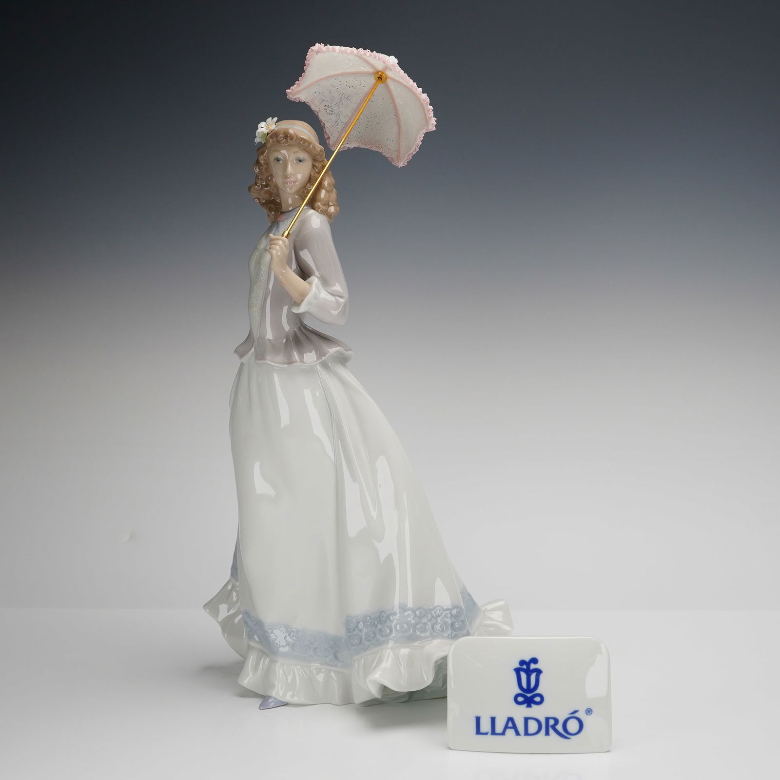 Lladro Porcelain Sculpture, On The Boulevard 01006545 with Plaque: The figure depicts a woman in turn-of-the-century style attire, consisting of a lavender jacket and a long white skirt with a blue patterned hem. The figure holds a removable porcelain parasol with a