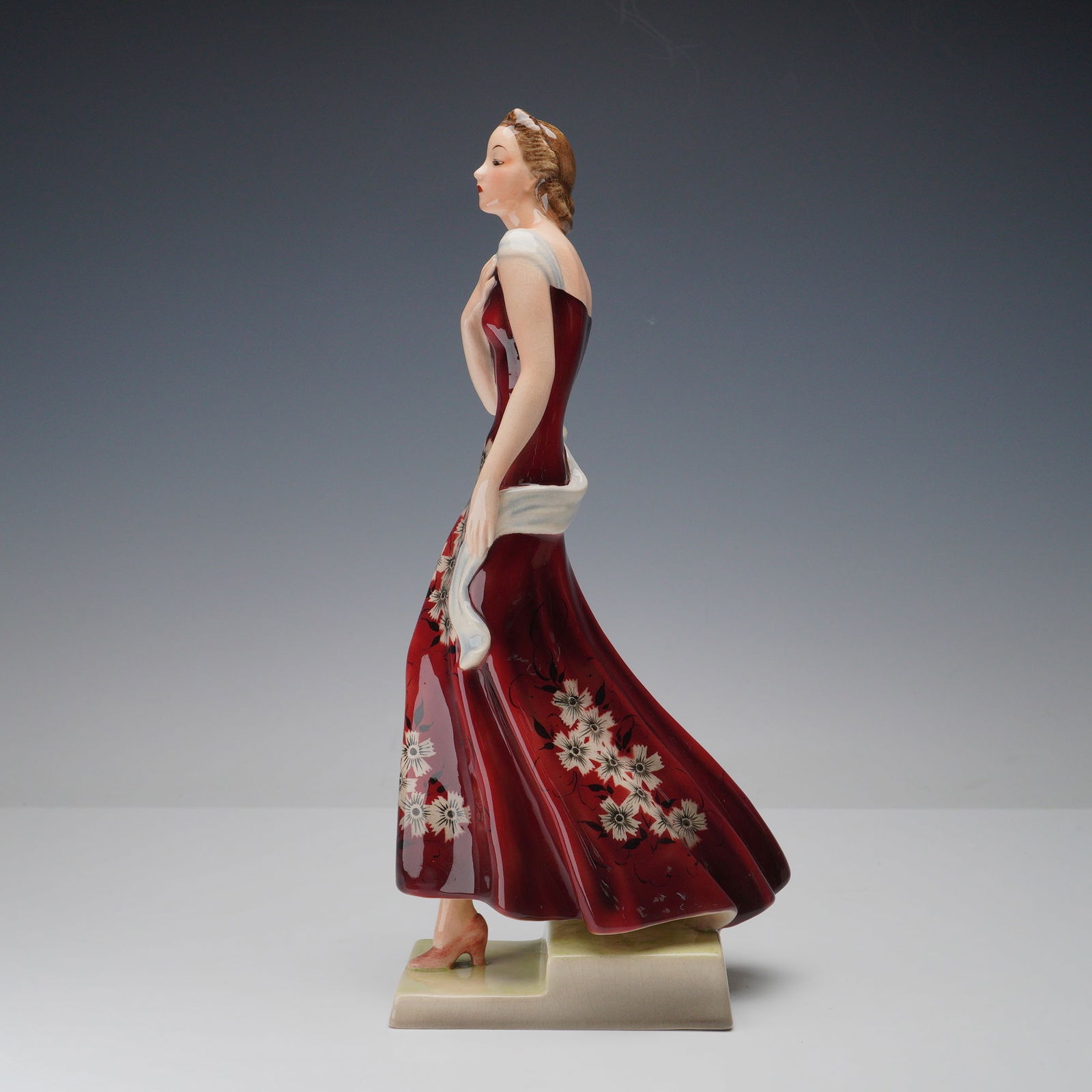 Goldscheider Woman in Floral Dress Porcelain Figurine: Striking Goldscheider porcelain figurine depicting a poised woman draped in a rich red gown adorned with floral motifs and accented by a pale shawl. The model captures the refined elegance and gracefu