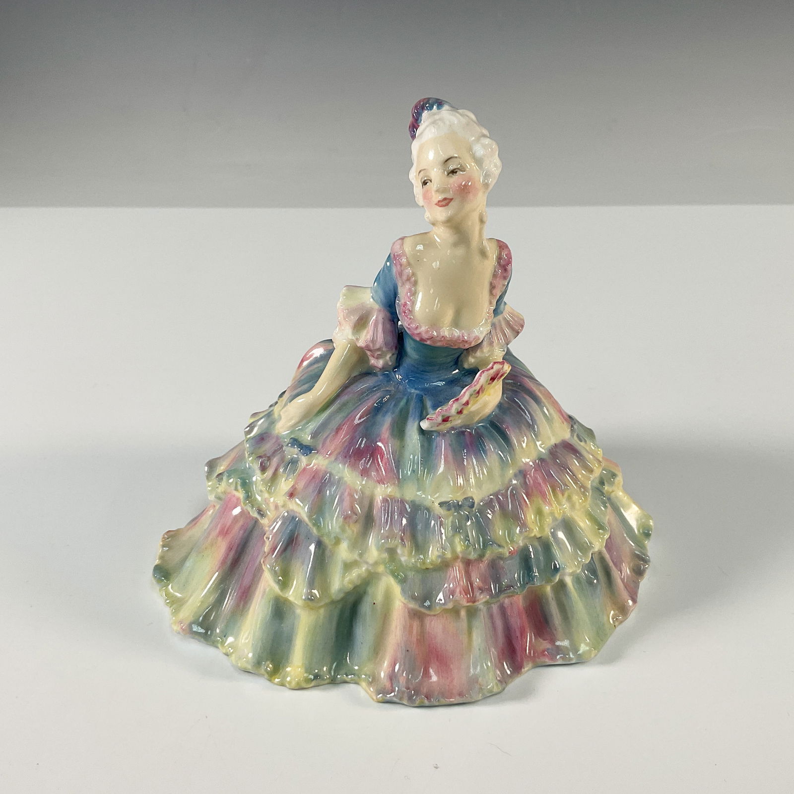 Lisette HN1524 - Royal Doulton Figurine: This Royal Doulton figurine is a charming creation designed by esteemed artist Leslie Harradine. Lisette showcases a woman in a flowing, ruffled gown painted in soft pastel shades of pink, blue, and y