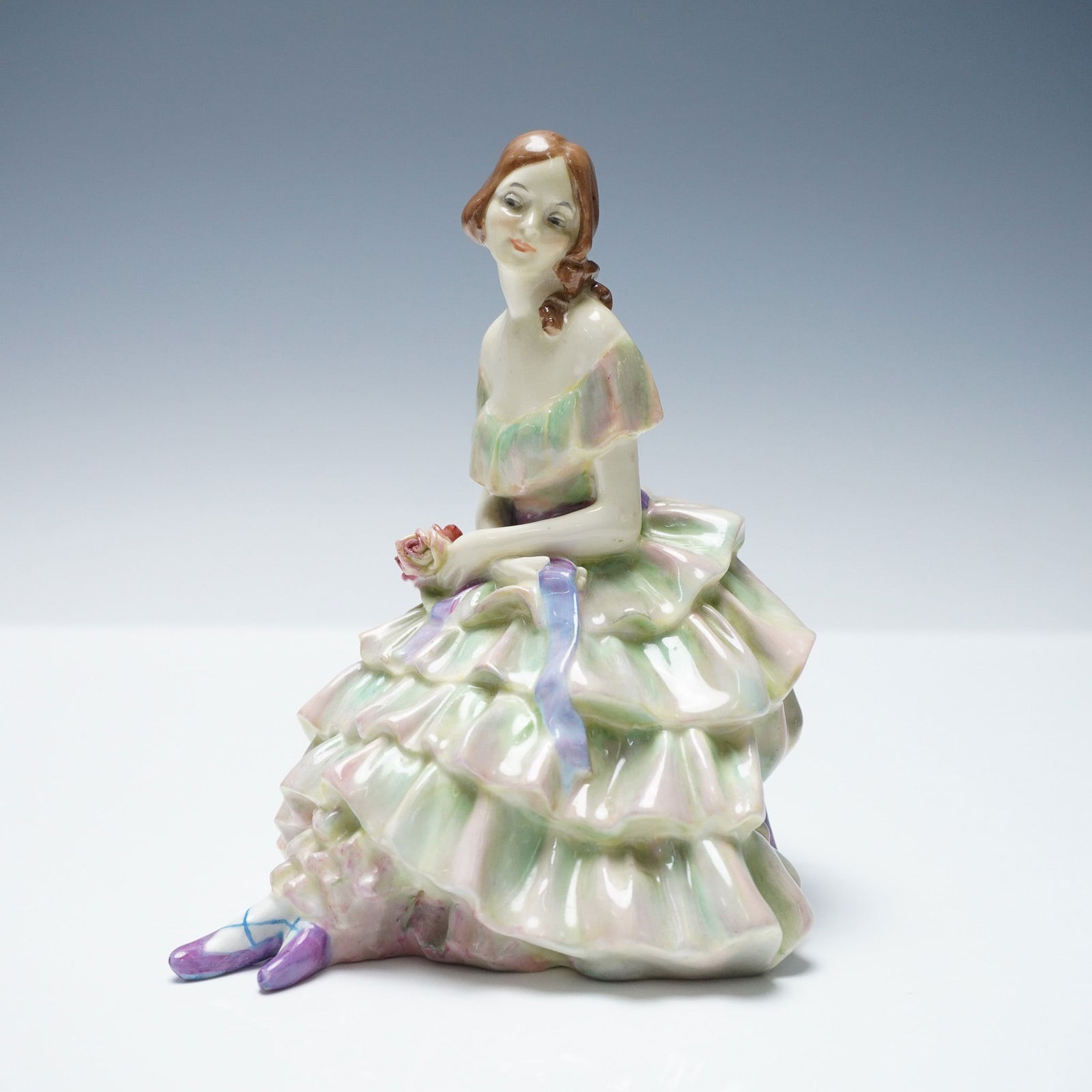 Royal Doulton Figurine, Gwendolen HN1494: The figure features a seated woman in a multi-tiered ruffled gown glazed in soft green and pink tones, with a lavender ribbon detail. She holds a flower in one hand and wears purple slippers. The base