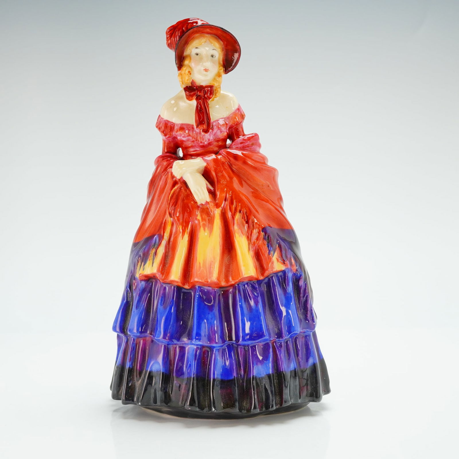 Royal Doulton Figurine, A Victorian Lady HN1277: Modeled woman in a full gown with layered skirts in bold red, orange, and violet tones, complemented by a matching bonnet tied with a ribbon. Royal Doulton backstamp. Artist: Leslie Harradine Dimensio