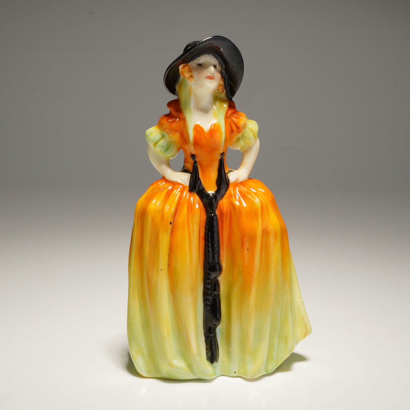Royal Doulton Miniature Figurine Patricia M8: A Patricia M8 Royal Doulton Figurine depicting a woman in an orange, yellow, and green gown and black hat, bearing impressed and handwritten Royal Doulton marks on the base. Dimensions: 2.50"L x 1.75"