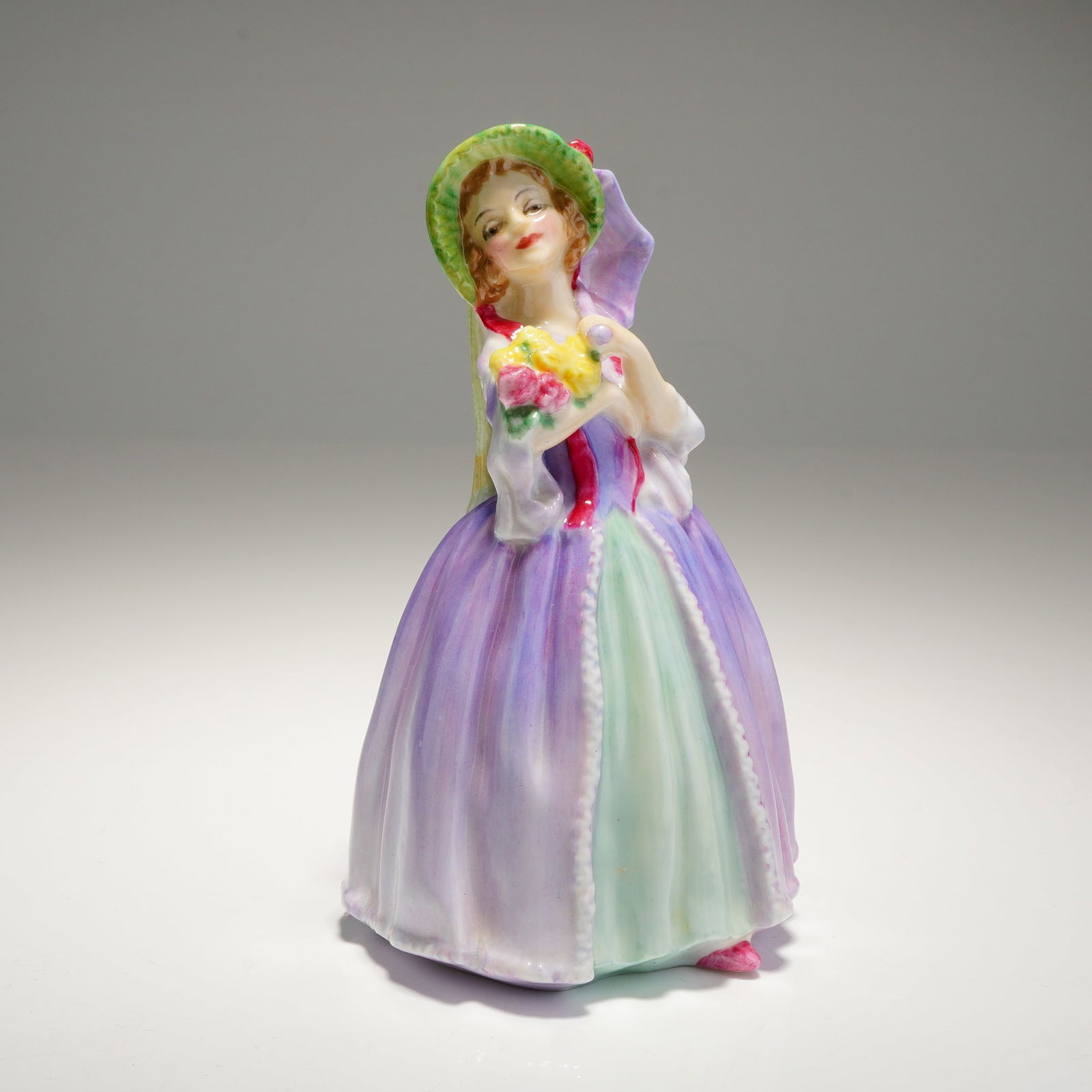 Royal Doulton Miniature Figurine June M71: A June M71 Royal Doulton Figurine depicting a lady in a purple, lilac, and mint green dress holding flowers and an umbrella, marked on the base. Dimensions: 2.50"L x 2.50"W x 4.25"H Weight: 3.40oz