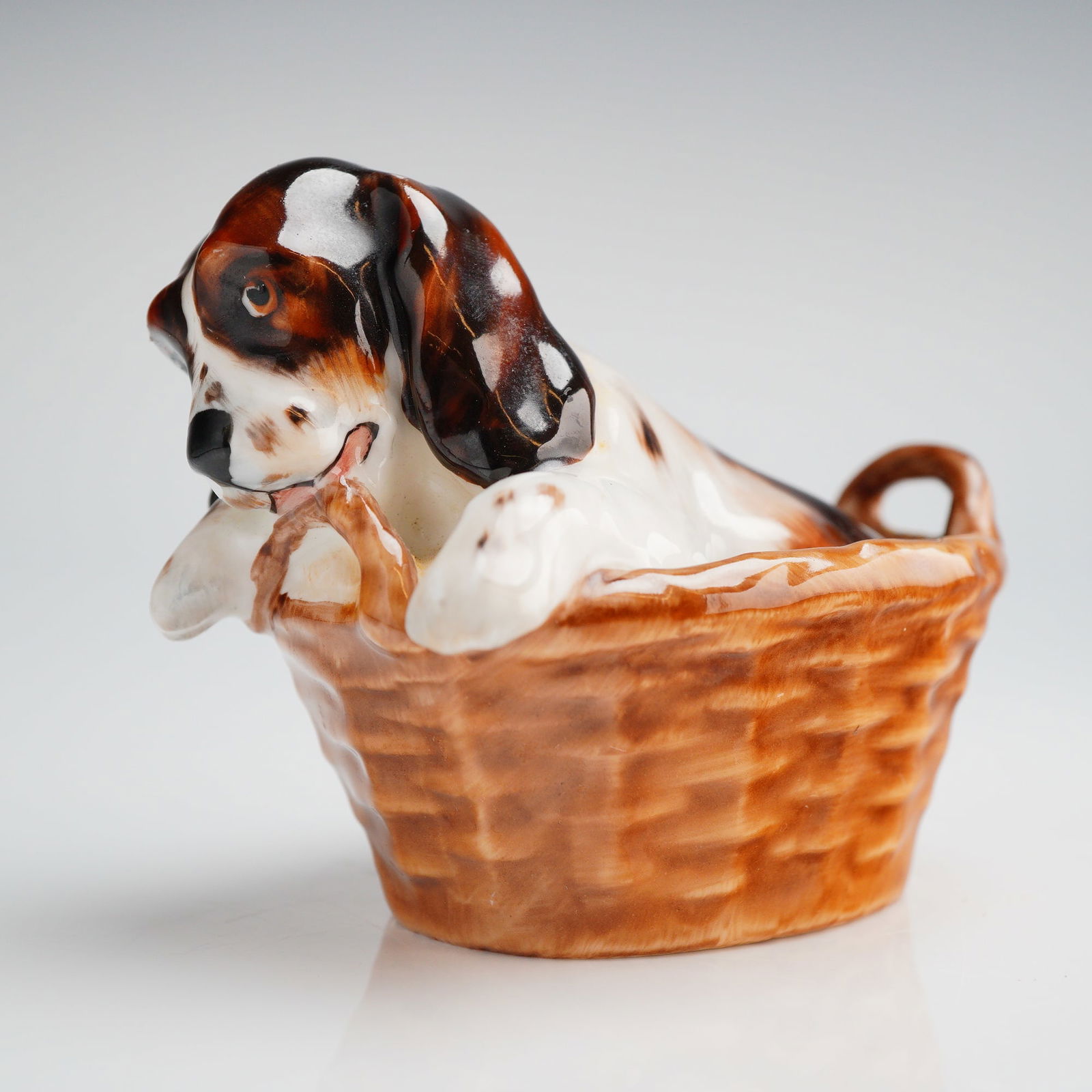 Royal Doulton Bone China Cocker Spaniel Puppy in Basket Figurine HN2586: A single glazed bone china figurine manufactured by Royal Doulton, depicting a Cocker Spaniel puppy playfully emerging from a woven basket. The dog features a tri-color coat of white, dark brown, and