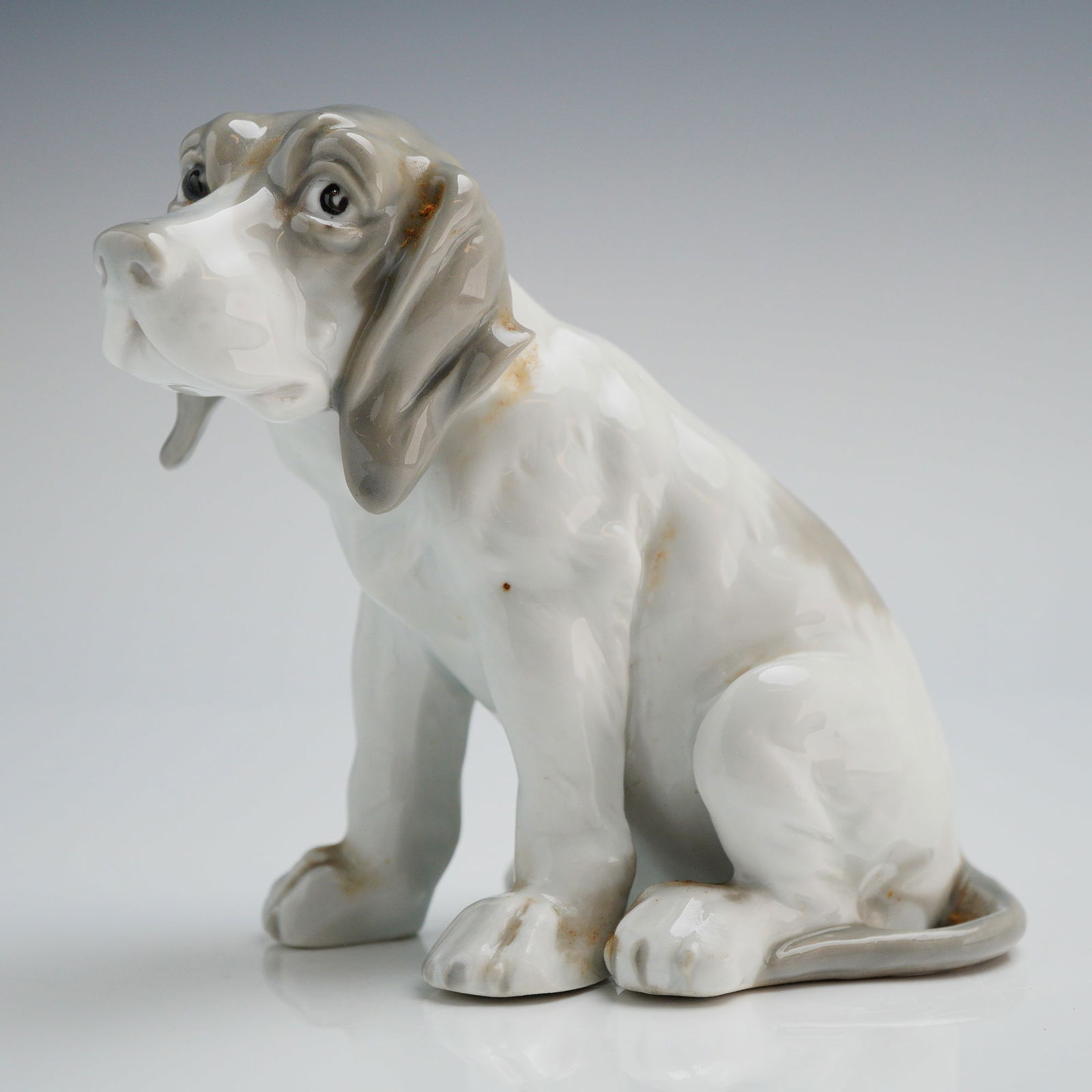 Gebruder Heubach Porcelain Figurine of a Seated Hound Puppy: A single glazed porcelain figurine by Gebruder Heubach, depicting a seated hound puppy, likely a Basset or Beagle. The figure is executed in a realistic style featuring a high-gloss white glaze. Hand-