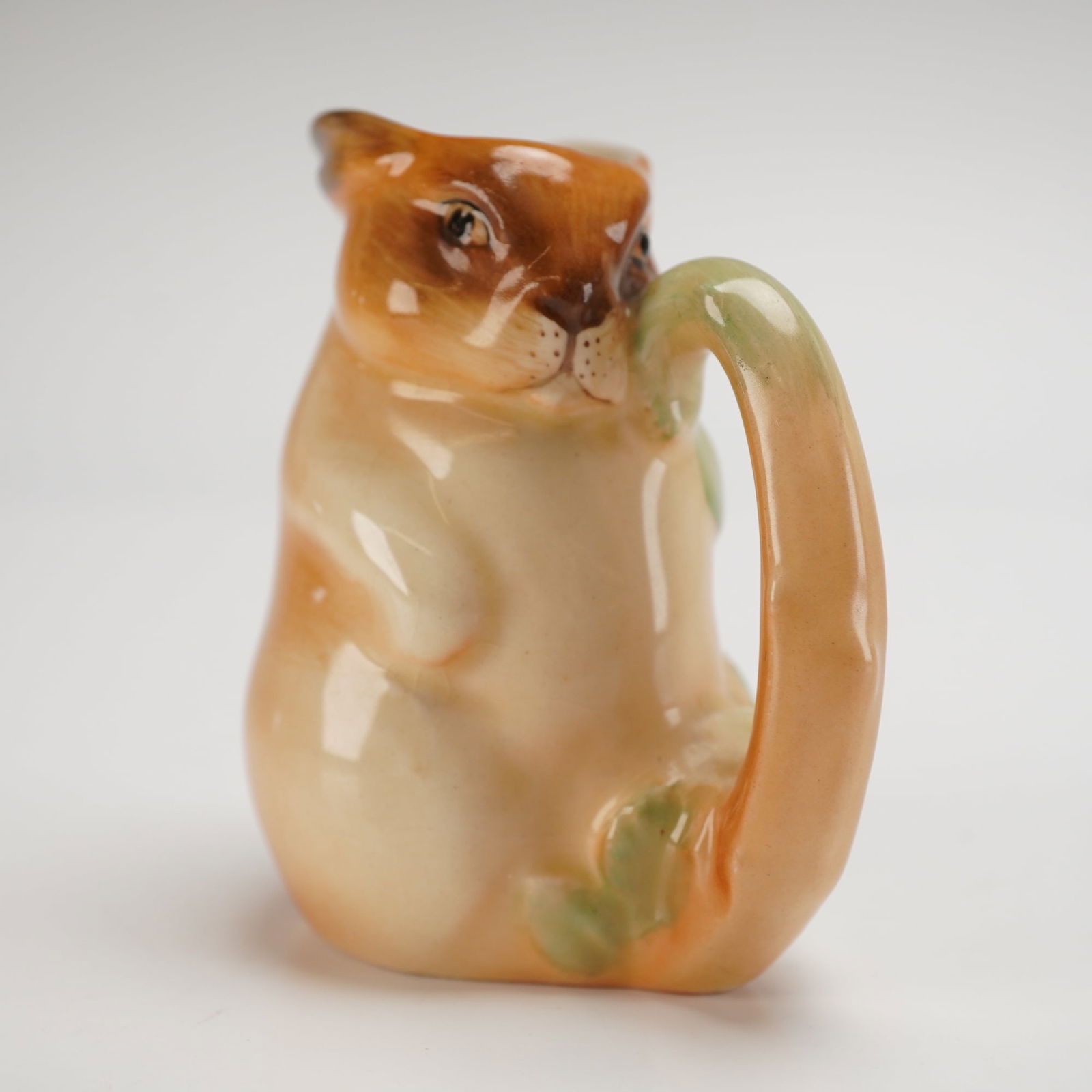 Royal Doulton Bunnykins Cream Jug, D6057: Part of the Bunnykins Breakfast Set. This charming piece is beautifully molded in the form of a whimsical rabbit, depicted in rich brown tones seated among vibrant green foliage. Crafted with the sign