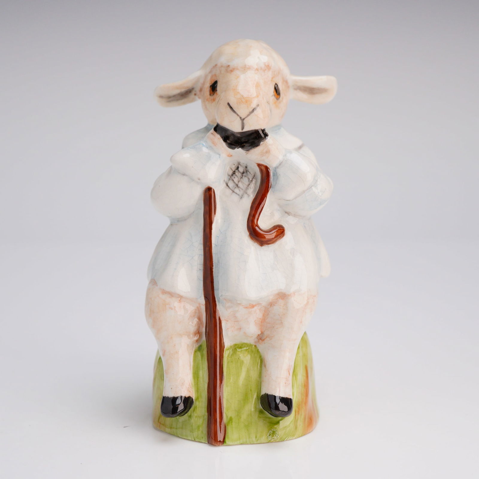 Royale Stratford Hand Painted Bone China Shepherd Sheep Figurine: A hand-painted glazed ceramic figurine by Royale Stratford, Staffordshire, England. The piece depicts an anthropomorphic lamb seated on a green base, holding a reddish-brown shepherd's crook. The lamb