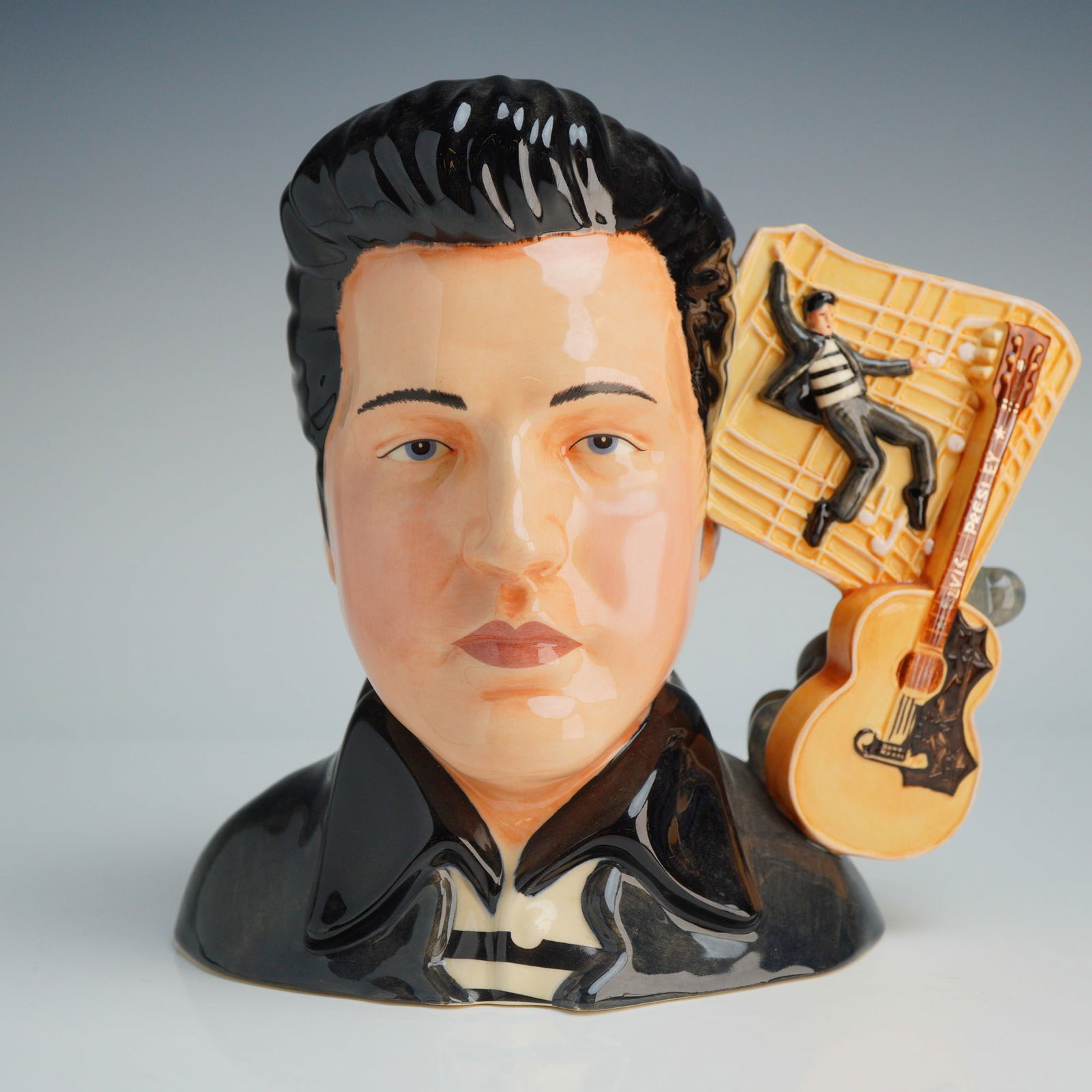 Royal Doulton Large Character Jug, Elvis Jailhouse Rock EP4: The piece features a glazed bust of the singer with a signature black pompadour hairstyle and a black collared shirt. The decorative handle is composed of an acoustic guitar inscribed with Elvis Presl