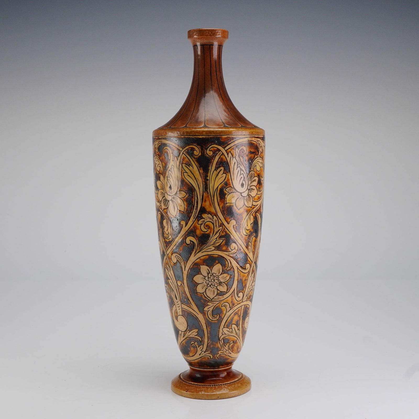 Martin Brothers Stoneware Vase: The vessel features a tall, slender, waisted cylindrical body rising from a flared circular foot to a narrow, ribbed neck and everted rim. The salt-glazed stoneware body is densely decorated with styl