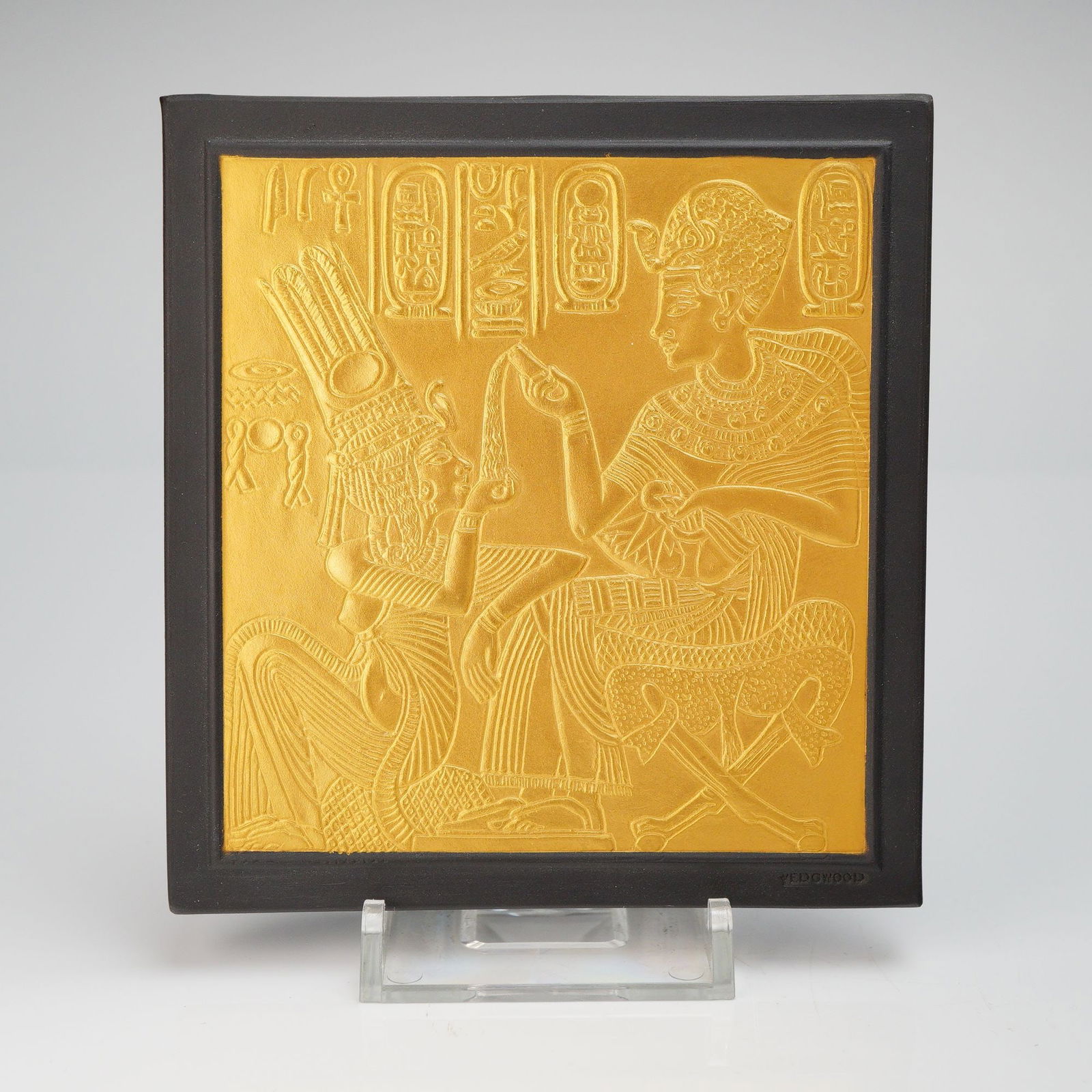 Wedgwood Gilded Black Basalt Egyptian Revival Plaque Enchantress: This square ceramic tile features a high-relief central panel finished in a vibrant gold-lustre, depicting the pharaonic scene of Tutankhamun and Ankhesenamun, a scene derived from the Golden Shrine.
