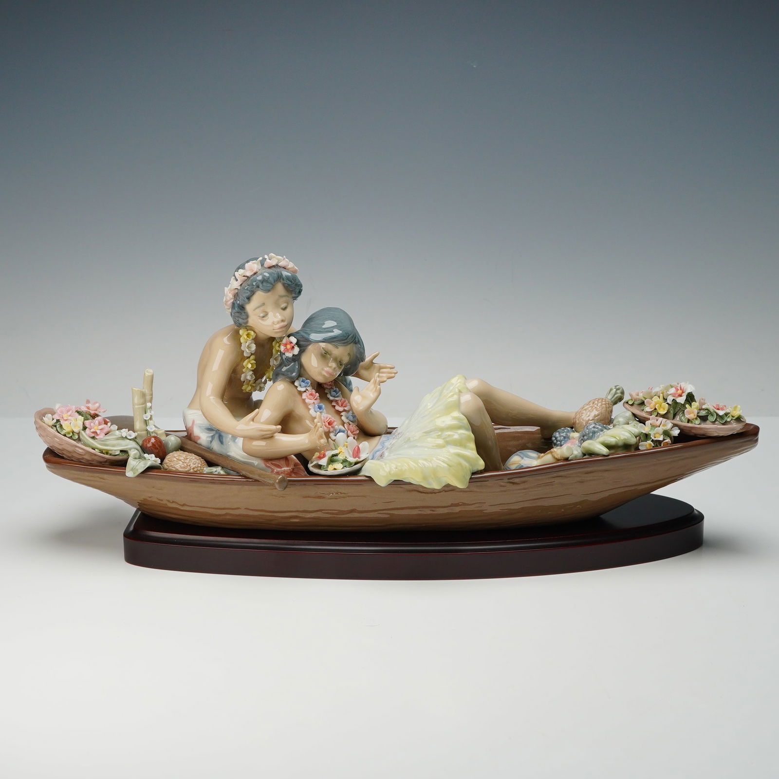 Lladro Figurine and Base, Lover's Paradise 1005779: A pair of island lovers rest together in a long wooden canoe, surrounded by flowers and fruit as they lean close in a relaxed embrace. Lladro backstamp. This item has its original box. Artist: Francis