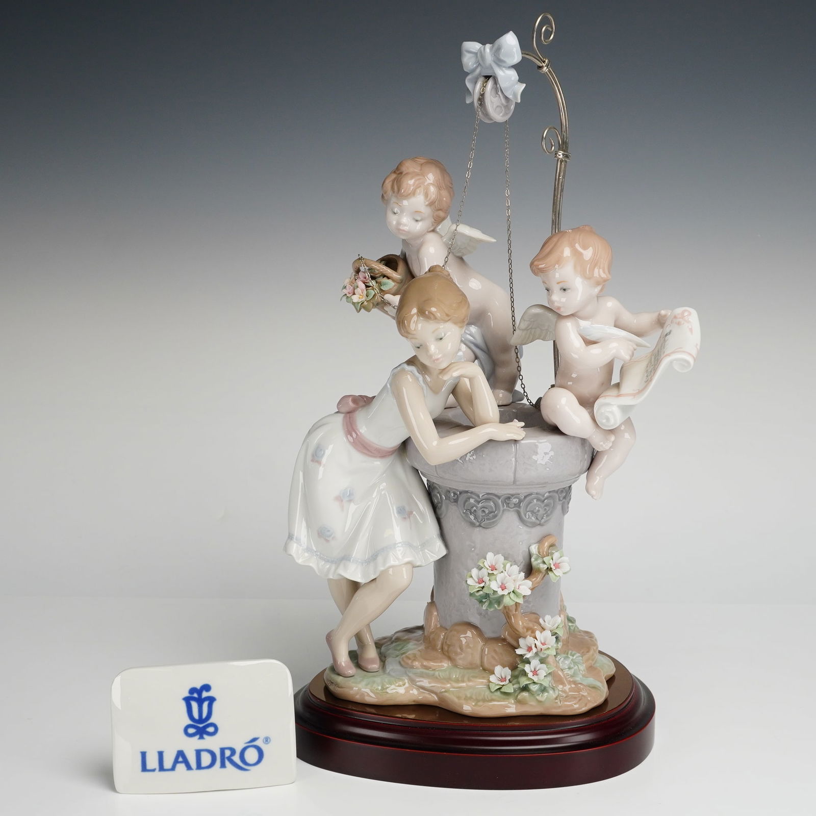 Lladro Porcelain Sculpture, May My Wish Come True 01001882 with Base: This limited-edition, 219 of 2000, sculpture depicts a young woman in a floral dress leaning against a decorative wishing well while accompanied by two winged cherubs. The piece features a metal arch