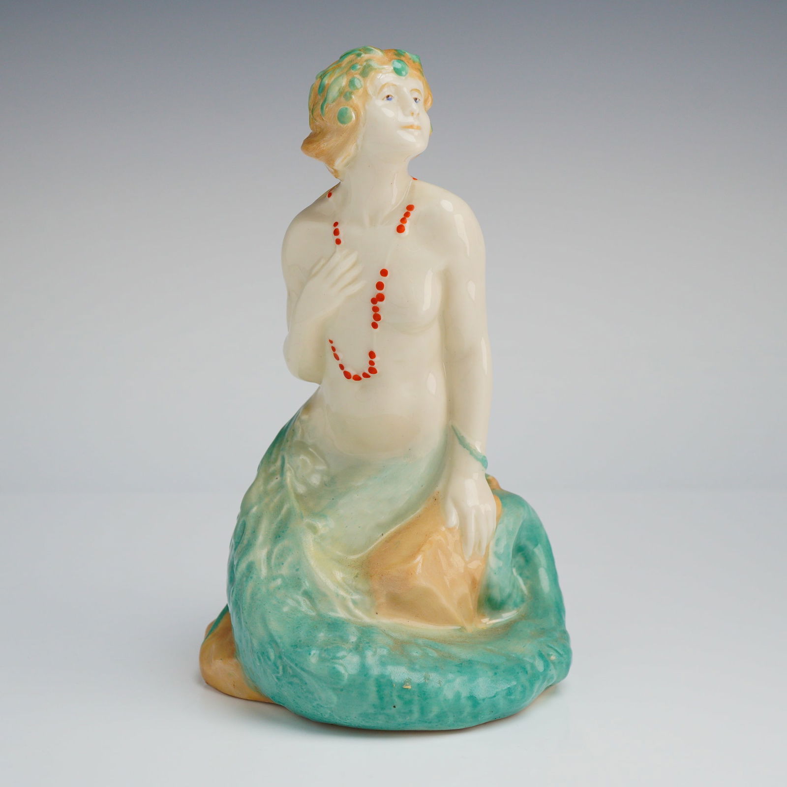 Royal Doulton Mermaid Figurine HN97: The piece is executed in an early 20th-century Art Deco style, depicting a siren or mermaid figure seated upon an ochre-colored rock formation. The upper torso is modeled nude and painted in flesh ton