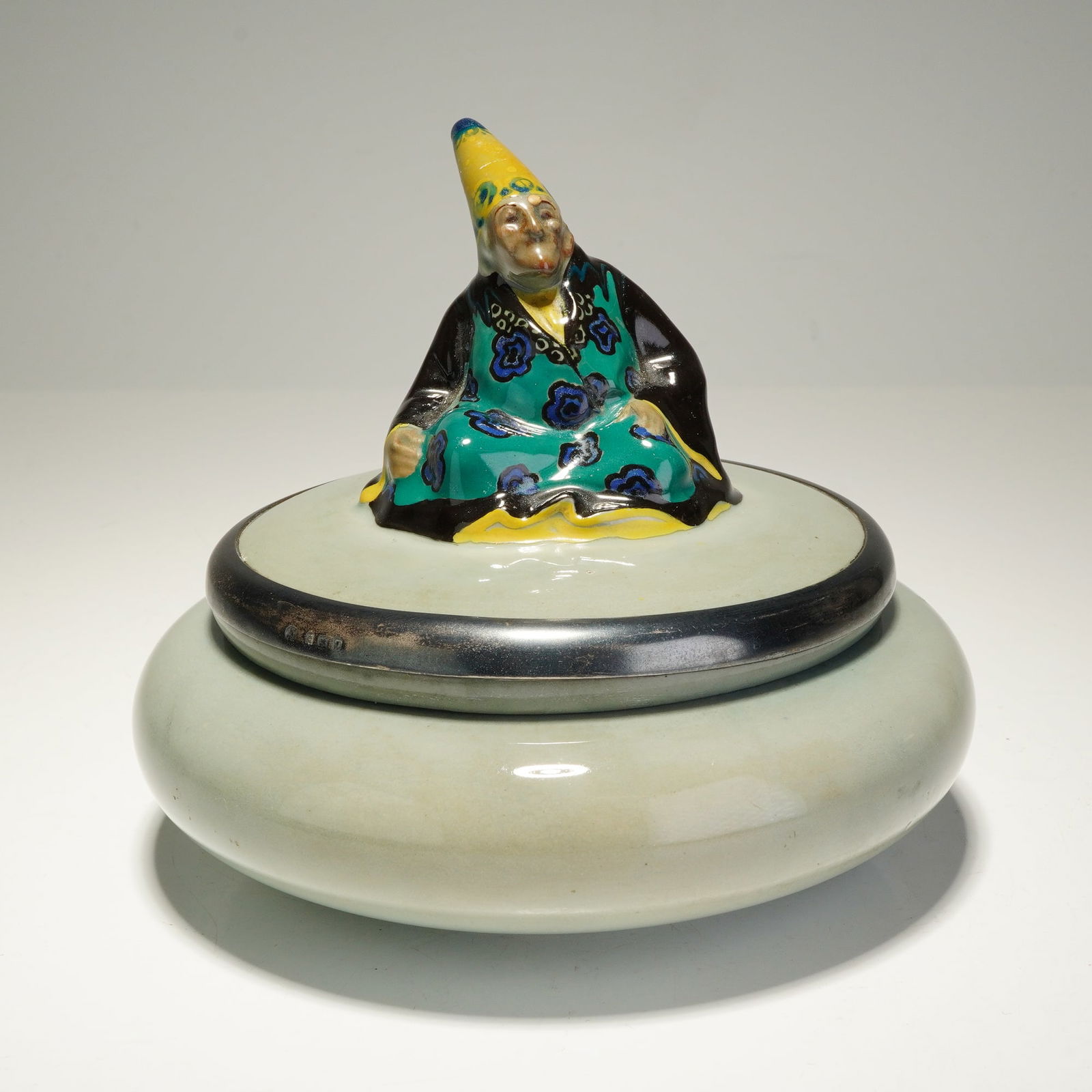 Royal Doulton Titanian and Sterling Silver Seated Figure Lidded Box: A rare English Titanian box surmounted by a lid modeled with a seated Mandarin-style figure, dressed in a black cloak over a green and blue floral tunic and wearing an yellow conical hat. The lid is f