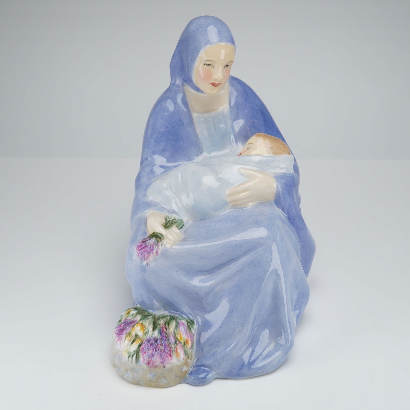 Royal Doulton Figurine, The Madonna of the Square, HN 1969: The figurine depicts the seated Virgin Mary holding the swaddled Christ Child, executed in a mid-20th-century figurative style. The Virgin is cloaked in a flowing periwinkle blue robe. A small basket