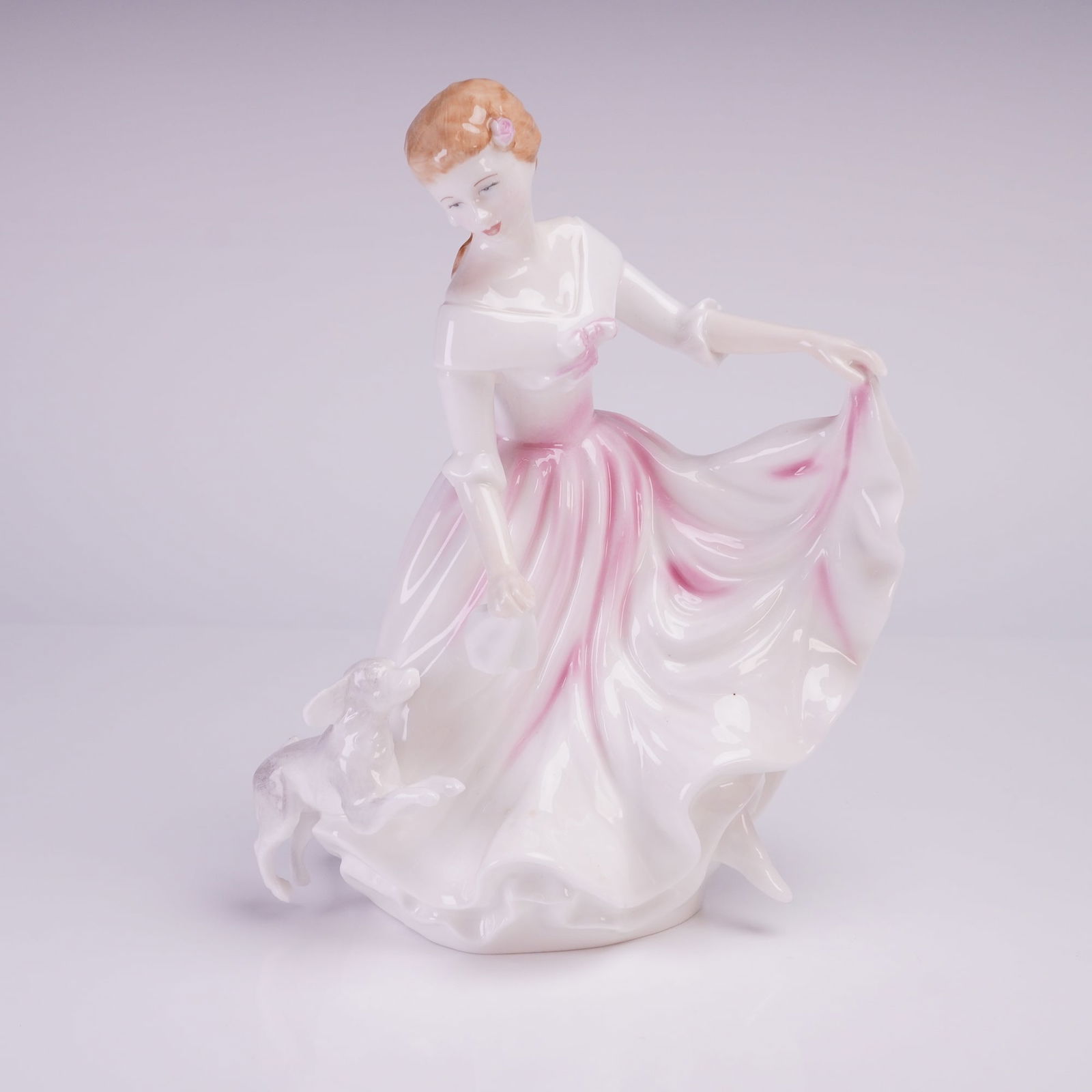 Royal Doulton Colorway Figurine, My Best Friend: The figurine, designed by Peter Gee, depicts a standing woman in a flowing white dress accented with pink coloration, captured in a dynamic pose lifting the skirt of her dress. A small white dog is po