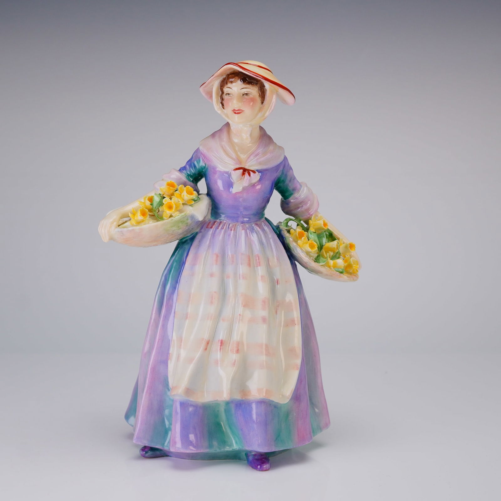 Royal Doulton Figurine Daffy Down Dilly HN1713: A Royal Doulton depicting a woman in a multi-colored purple and teal striped dress holding two baskets of yellow flowers, marked underneath with the brand logo and registration number Rd N803697. Dime