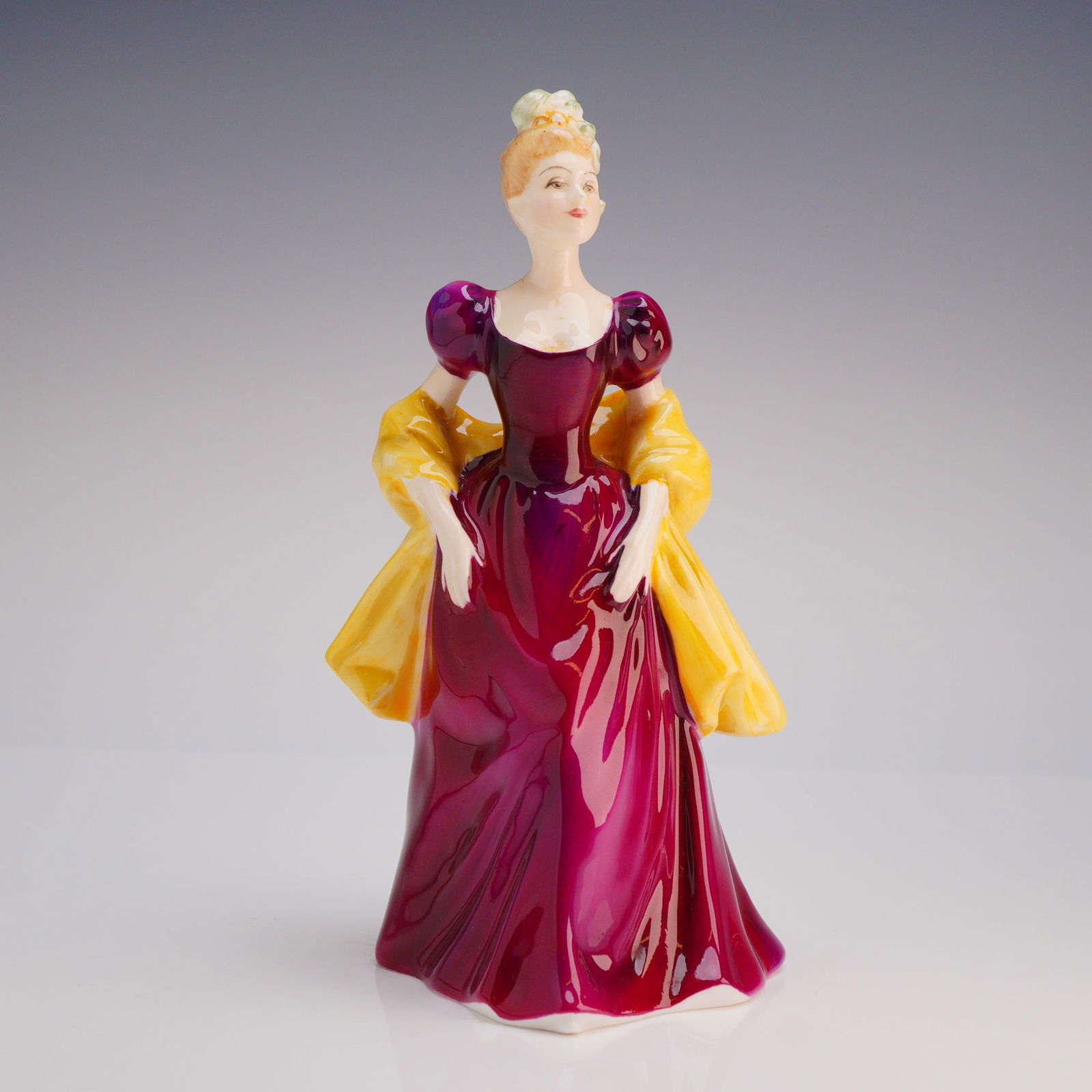 Royal Doulton Figurine Loretta HN 2337: The figure is styled in a classical 20th-century decorative manner, depicting a standing female figure dressed in a voluminous, high-gloss magenta gown with puffed sleeves, accented by a bright yellow