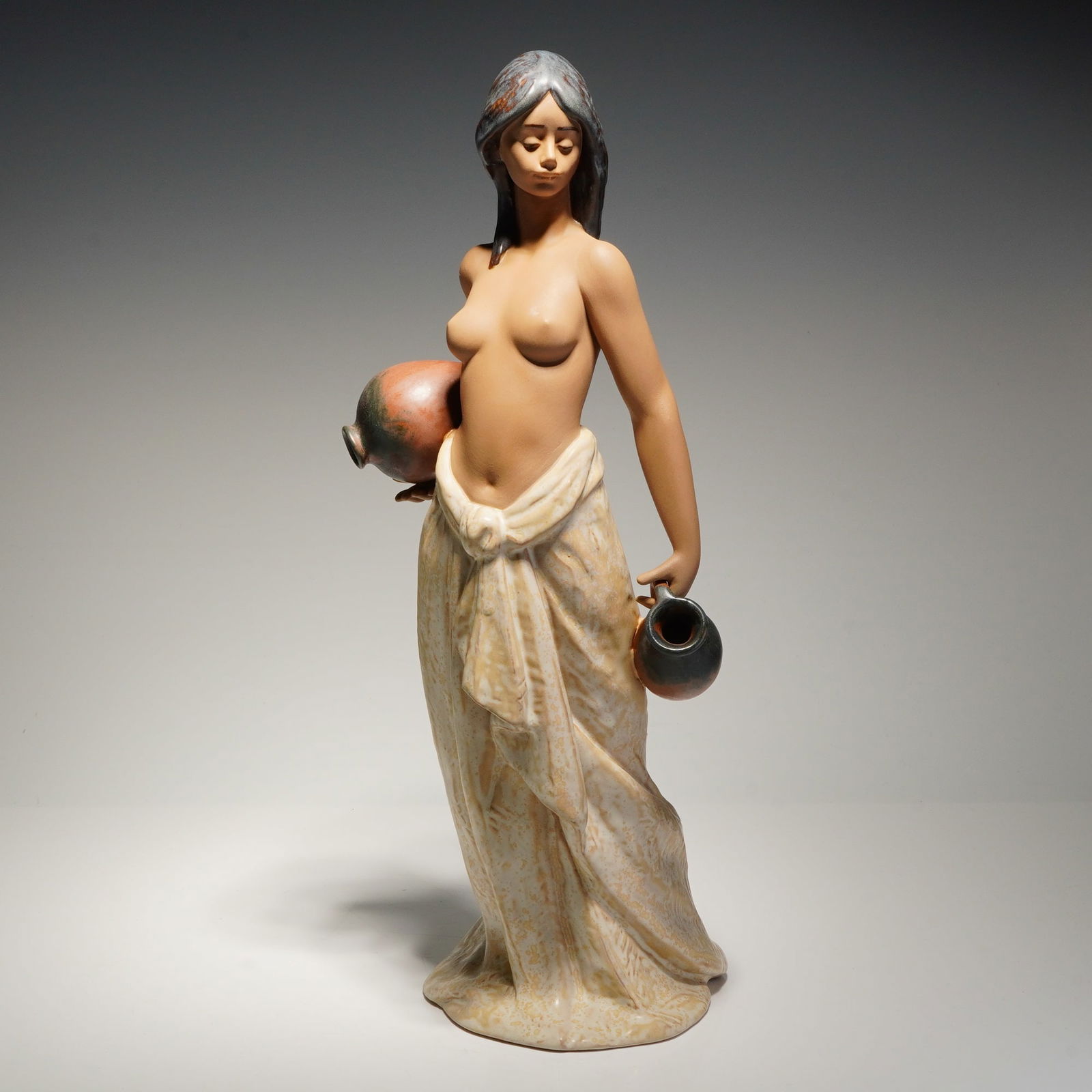 Lladro Porcelain Sculpture Water Girl 01012323: This Lladro piece depicts a semi-nude female figure carrying two water jugs. The sculpture is rendered in a matte gres finish with a palette consisting of muted brown, gray, and terracotta tones. The
