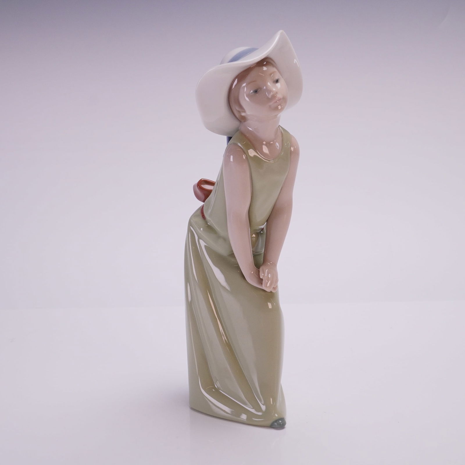 Lladro Porcelain Figurine, Curious Girl with Straw Hat 01005009: This figurine is a glazed porcelain designed in the manufacturer's characteristic slender style. The figure is depicted in a floor-length, sleeveless olive-green dress featuring a large brownish-red b