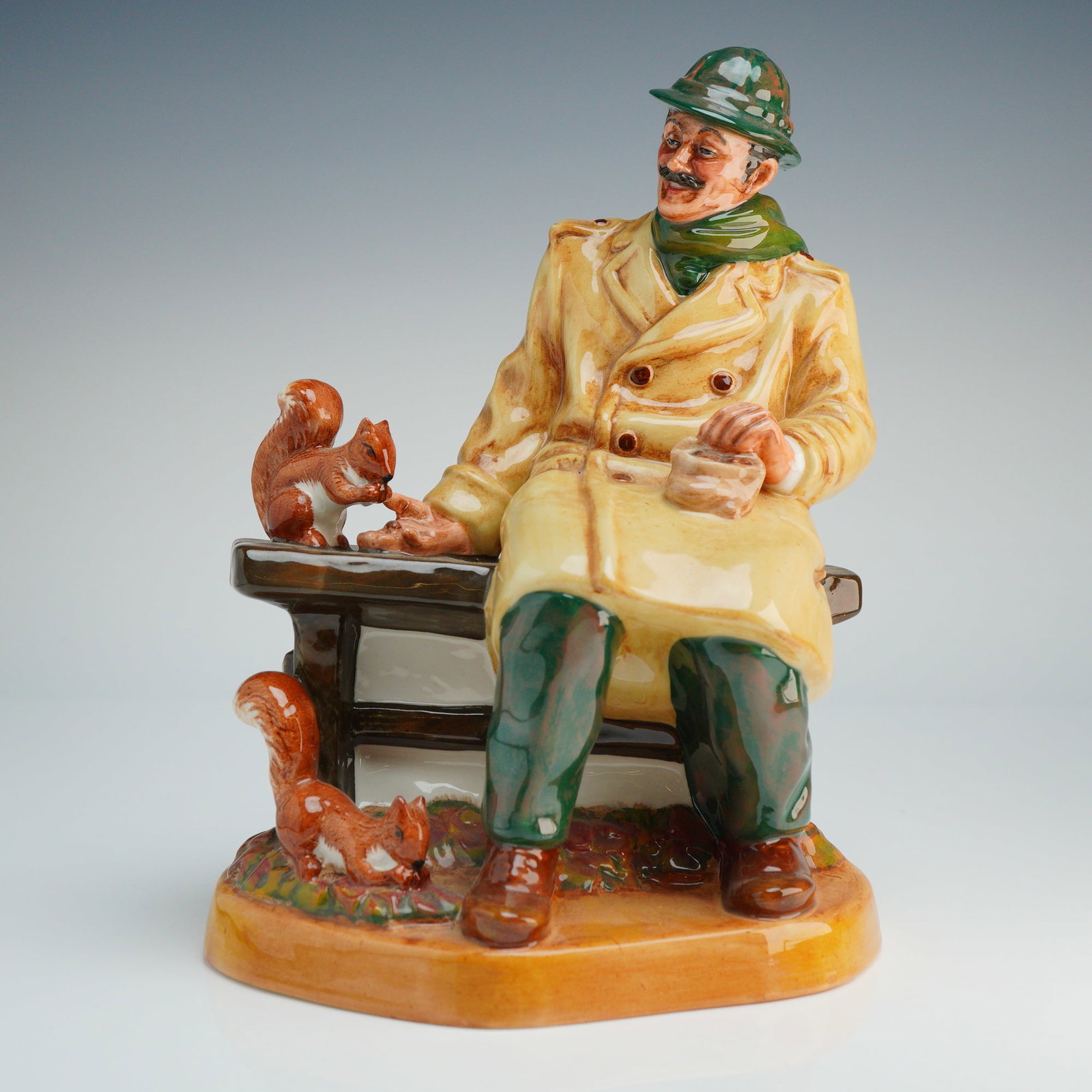 Royal Doulton Figurine, Lunchtime HN2485: This polychrome figure is finished with a high-gloss glaze and depicts a seated man with a mustache wearing a green fedora-style hat, a green scarf, and a long beige double-breasted trench coat. The s