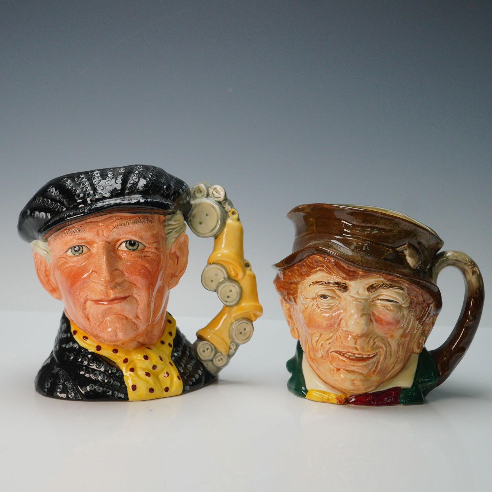 2pc Royal Doulton Character Jug Set Pearly King D6760 and Paddy: Pair of Royal Doulton character jugs, featuring the Pearly King D6760, hand made and hand decorated, modelled by Stanley James Taylor in 1986, with a distinctive button-adorned cap and scarf. Accompan