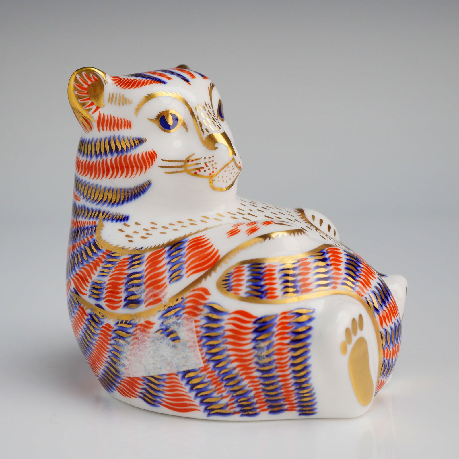 Royal Crown Derby Tiger Cub Paperweight: A Royal Crown Derby bone china paperweight depicting a seated tiger cub in the Imari style. The figurine is constructed with a white ground and decorated with orange and cobalt blue feather-like patte