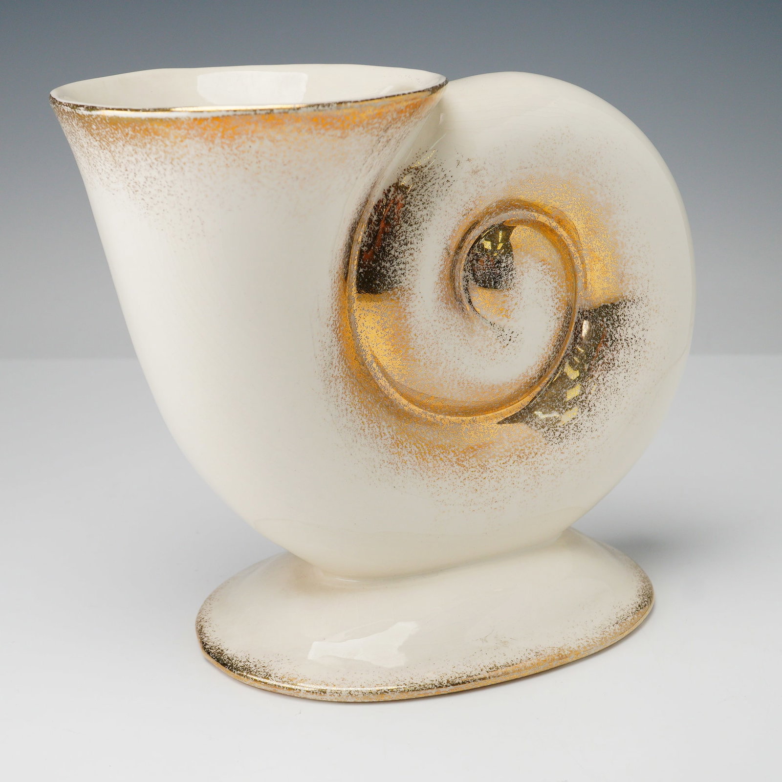 Pitt Petri Shell Form Footed Vase: A mid-century modern Pitt Petri vase crafted in the shape of a stylized shell, featuring a smooth cream glaze with shimmering gilt accents. The sculptural spiral form rises from an oval footed base, s