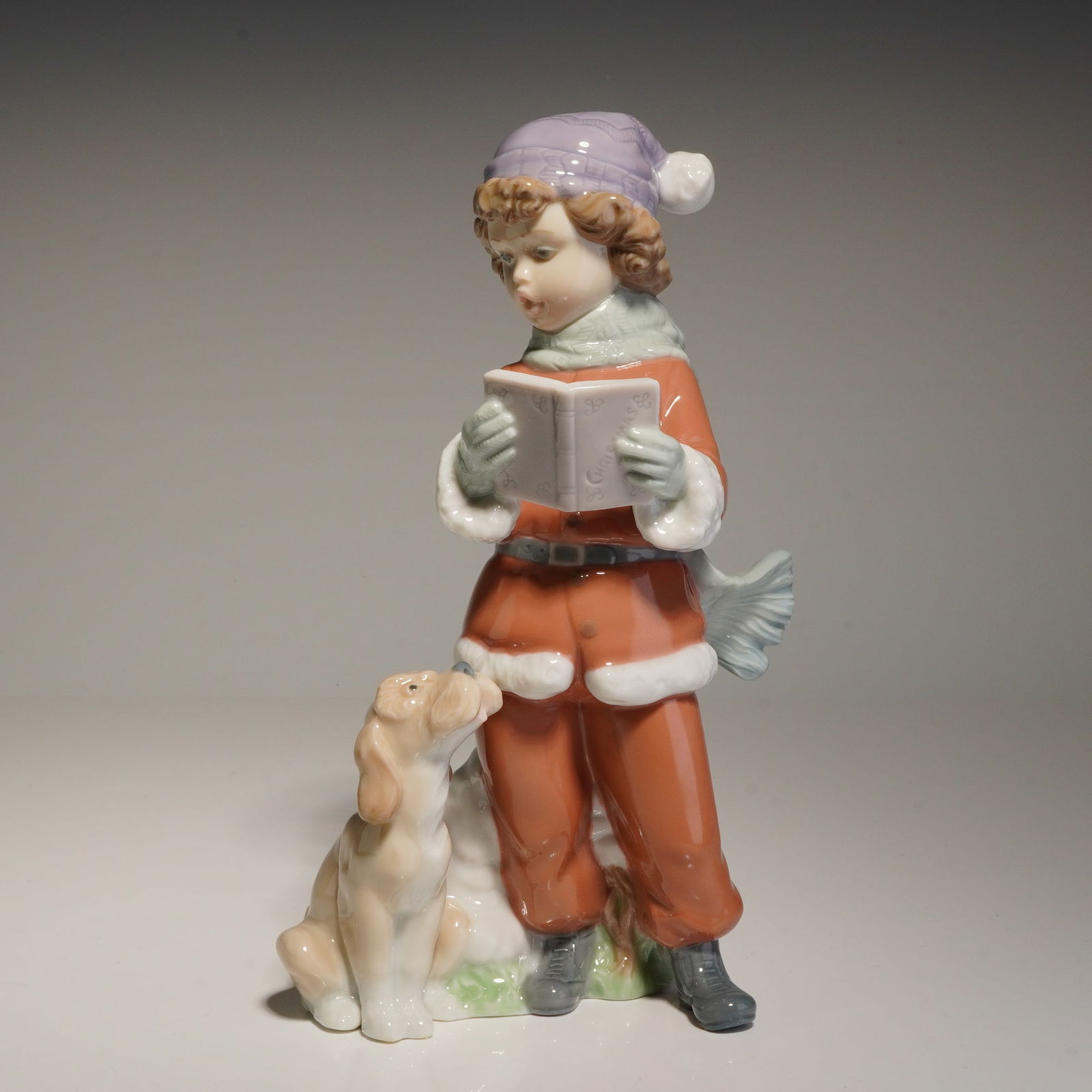 Lladro Porcelain Figurine, Christmas Duet 01006714: This figurine, produced by Lladro in Spain, depicts a young boy in a winter suit and purple cap reading from a book while a small dog sits at his side. The piece features the soft, muted palette and g
