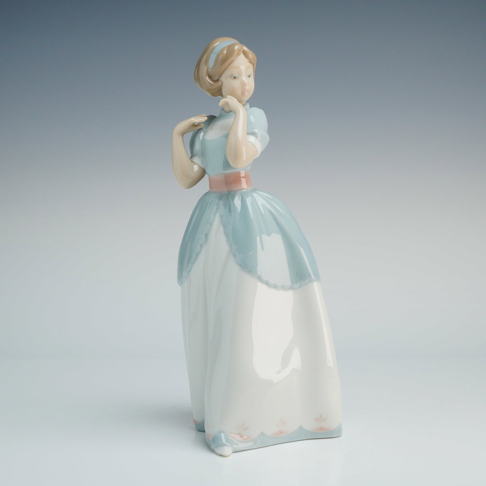 Lladro Porcelain Figurine, A Proper Pose 1006755: This glazed ceramic figure, hand-crafted in Valencia, Spain, depicts a young woman in a standing pose. She is dressed in a light blue bodice with puffed sleeves, a white full-length skirt featuring a