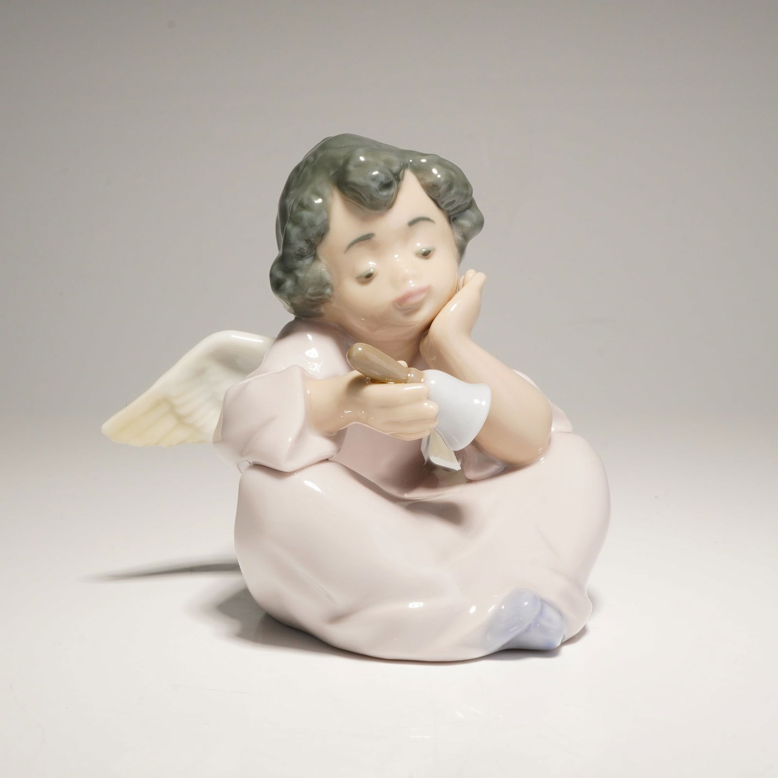 Lladro Porcelain Figurine, Heavenly Chimes 01005723: This glazed porcelain figure depicts a seated angel in a contemplative pose, resting its head on one hand while holding a small bell. The piece is finished in a glossy glaze with a soft pastel color p