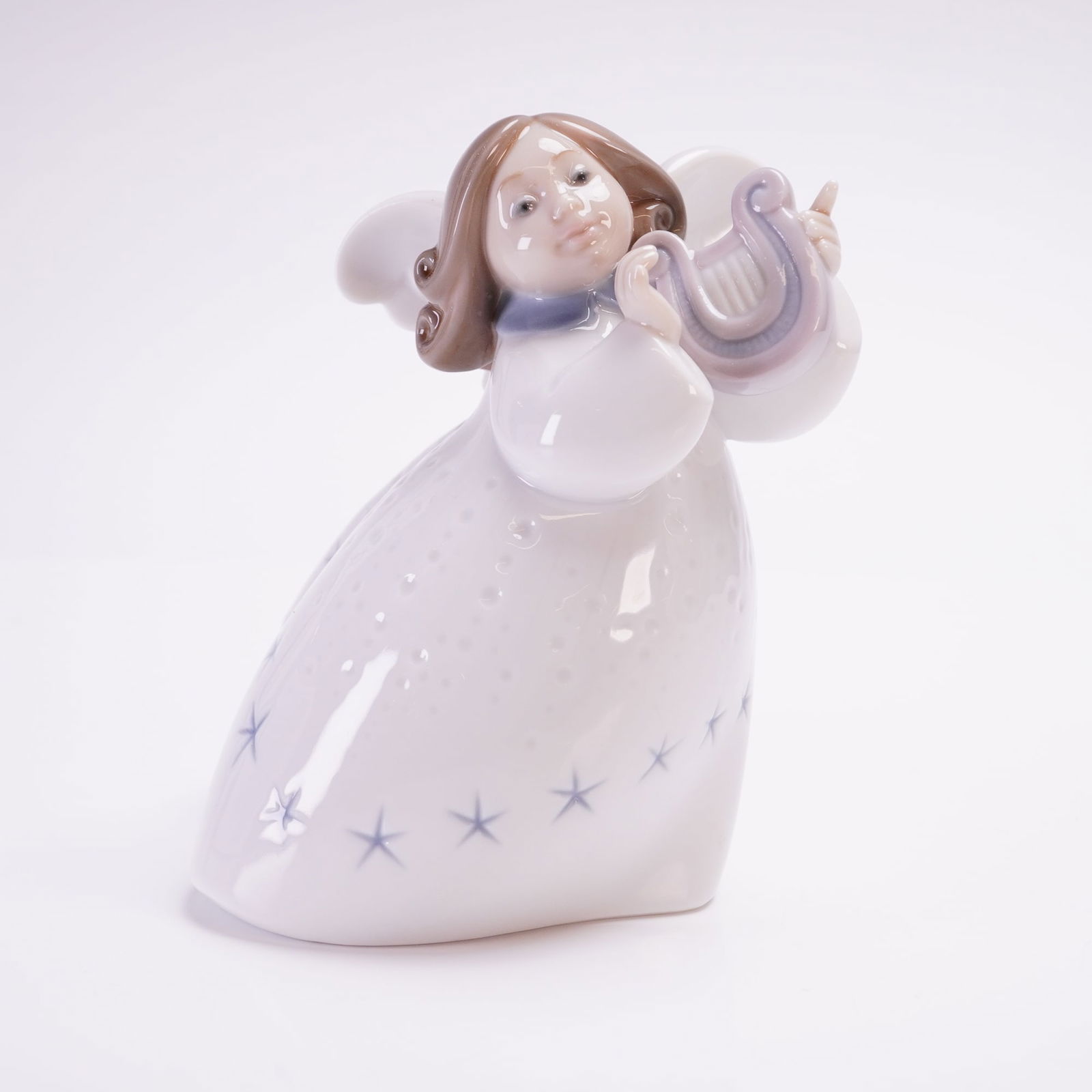Lladro Porcelain Figurine, Little Angel with Lyre 01006528: This Lladro depicts a winged figure playing a musical instrument. The figure is dressed in a flowing white gown accented with light blue stars and finished in a characteristic high-gloss pastel palett