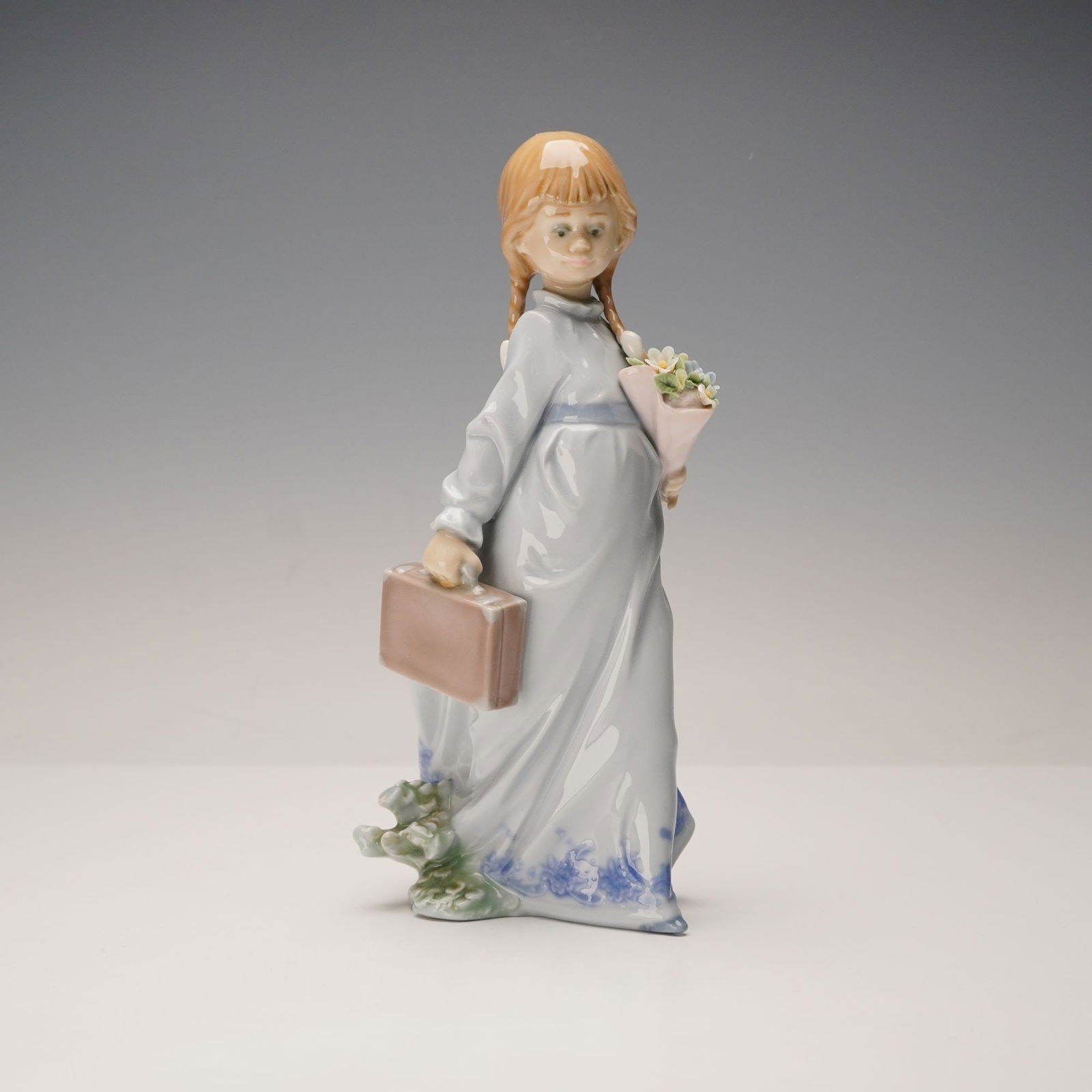 Lladro Porcelain Figurine, School Days 1007604: This glazed porcelain figurine depicts a young girl with braided hair, dressed in a flowing light blue gown with a paler blue sash and blue floral details at the hem. She carries a rectangular suitcas