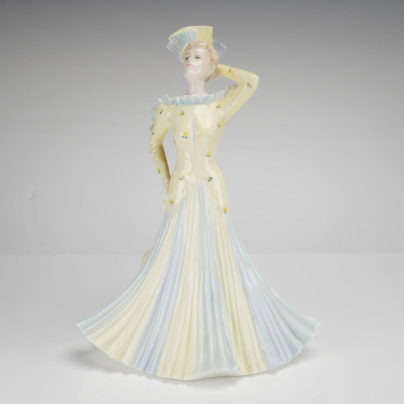 Coalport David Shilling Collection Figurine, L.A. Gala: The figure is modeled in a pale-yellow bodice with small floral accents and a ruffled neckline, leading into a wide, pleated skirt with alternating pale yellow and light blue panels. The figure is pos