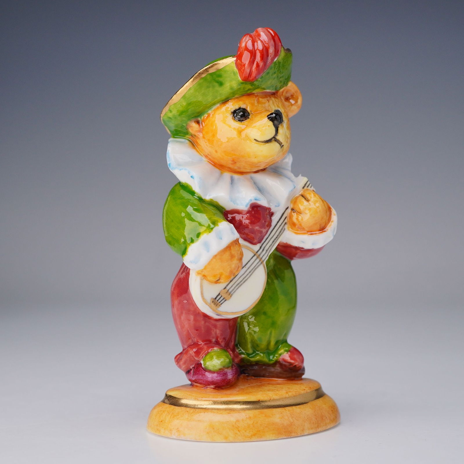 Halcyon Days Porcelain Figurine, Bear with Banjo: Hand-painted porcelain bear figurine wearing a green hat and patterned garment, depicted playing a banjo. Presented in its original Halcyon Days fitted satin-lined box. A charming and whimsical exampl