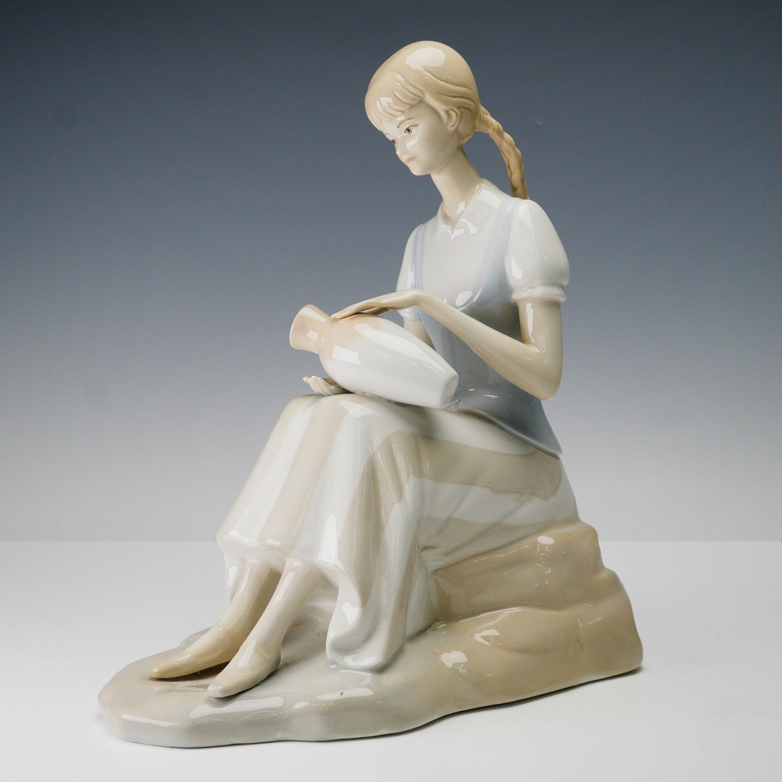 Porceval Spain Porcelain Figurine, Girl with Vase: The figure is crafted in soft pastel tones with a glossy glaze, highlighting delicate details such as the girl's braided hair, serene expression, and flowing dress. She is shown resting on a naturalis