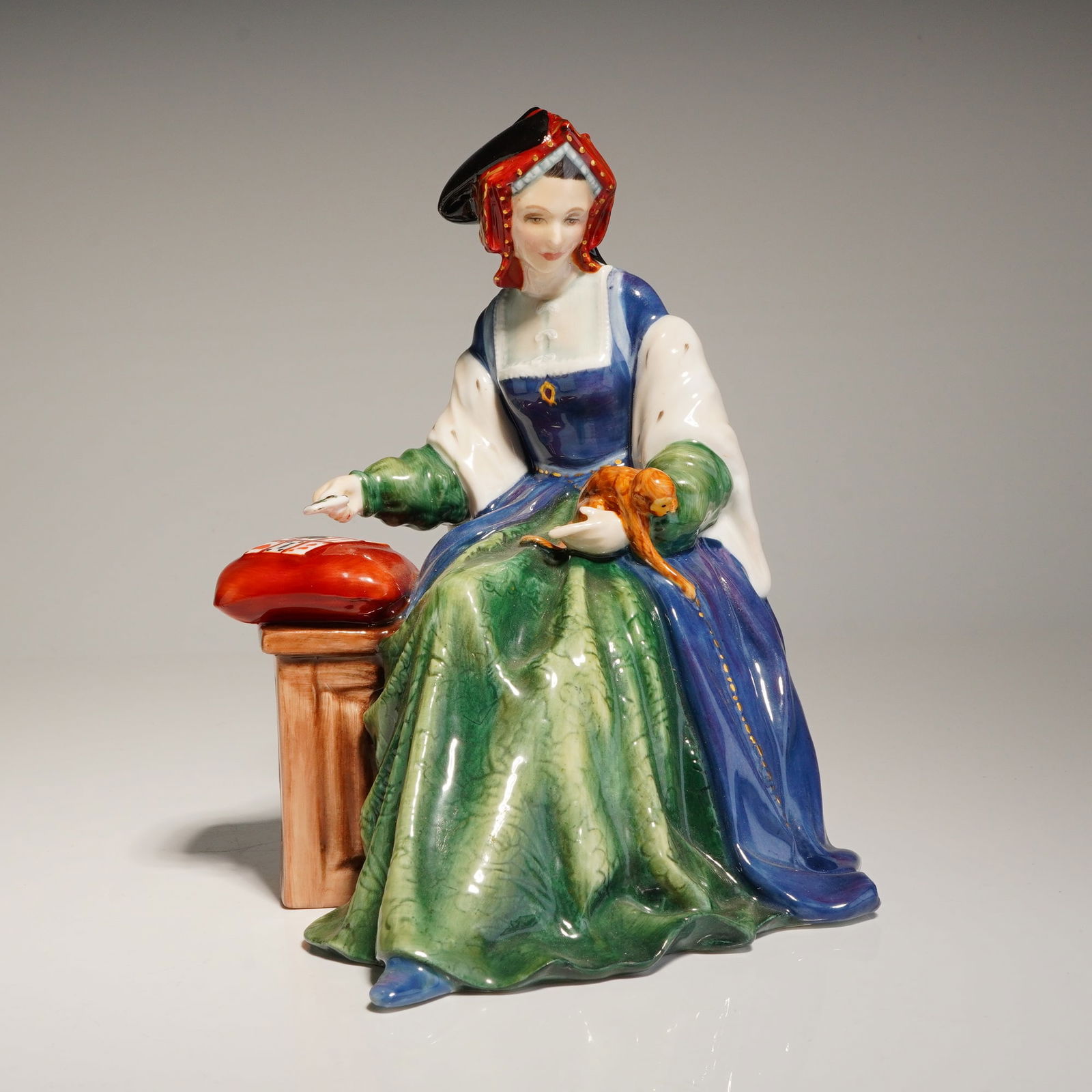 Royal Doulton Figurine, Catherine Aragon HN3233: A Royal Doulton glazed porcelain is from the Tudor Queens series. The hand-painted figure depicts the subject seated in a gown of blue and green with white sleeves and a gable hood. The figure is show