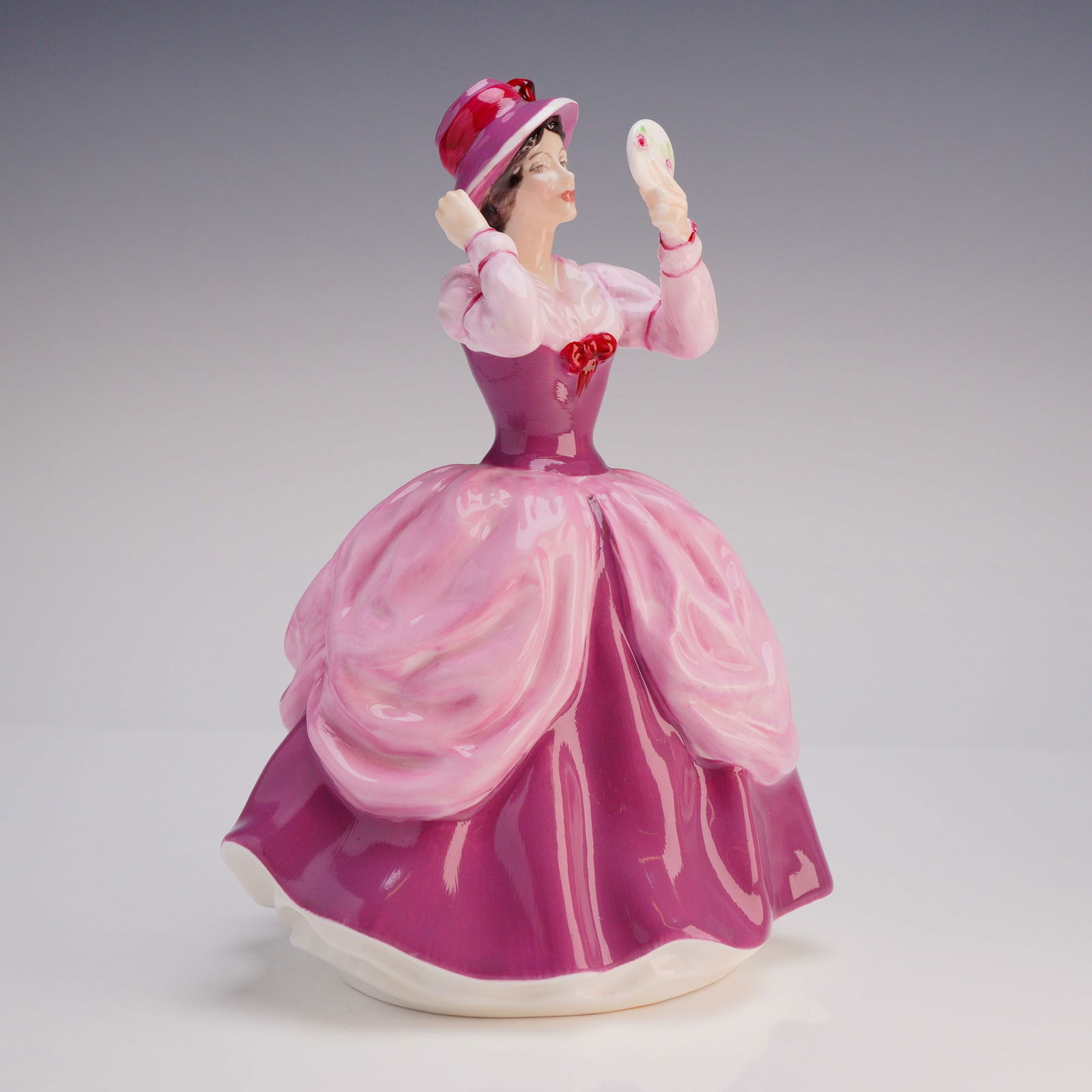 Royal Doulton Figurine Lady Pamela HN 2718: The piece depicts a woman in a voluminous period gown and wide-brimmed hat, checking her reflection in a small hand mirror accessory. The figurine is hand-painted primarily in shades of pink and magen