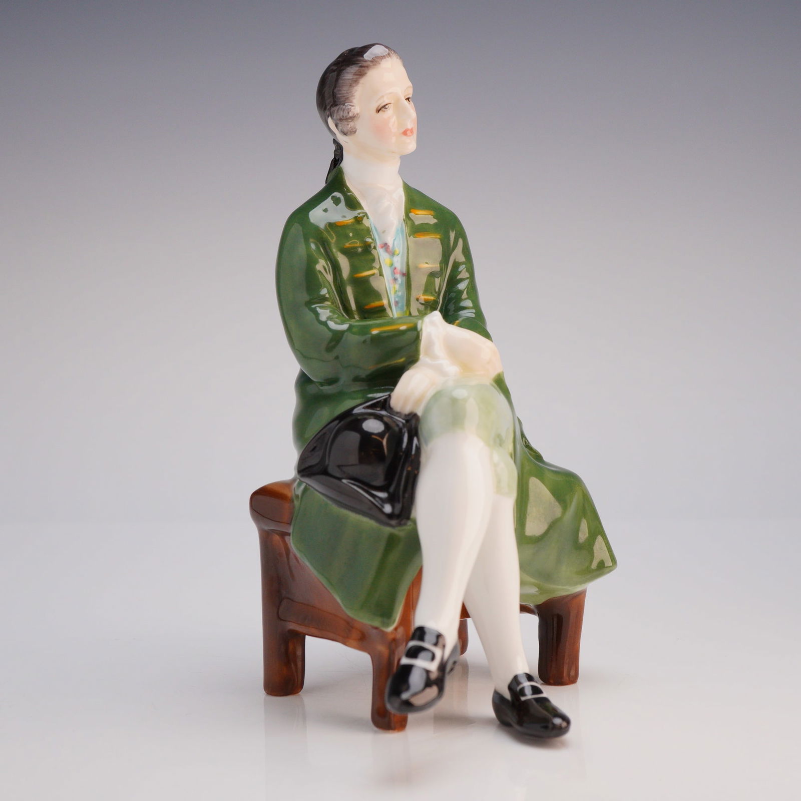 Royal Doulton Figurine A Gentleman from Williamsburg HN 2227: The figure depicts a gentleman styled in 18th-century attire, seated on a low brown stool. The gentleman is wearing a highly glossy dark green long coat over lighter patterned clothing, white breeches