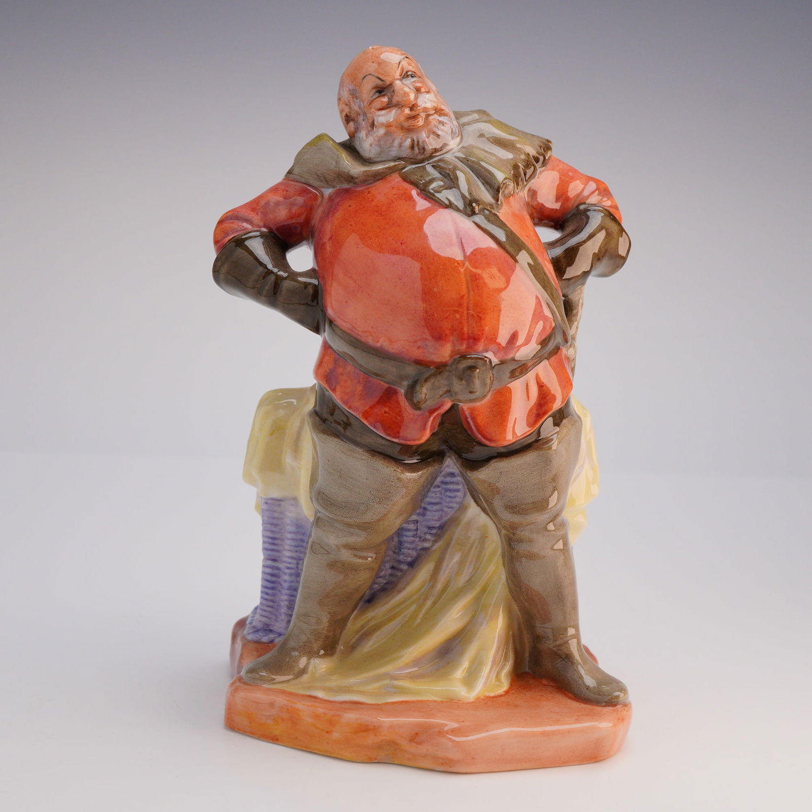 Royal Doulton Figurine Falstaff HN 2054: The figurine depicts the stout male character from Shakespeare, portrayed with a white beard, stern expression, and hands placed on his hips. He is dressed in historical attire featuring a vibrant ora
