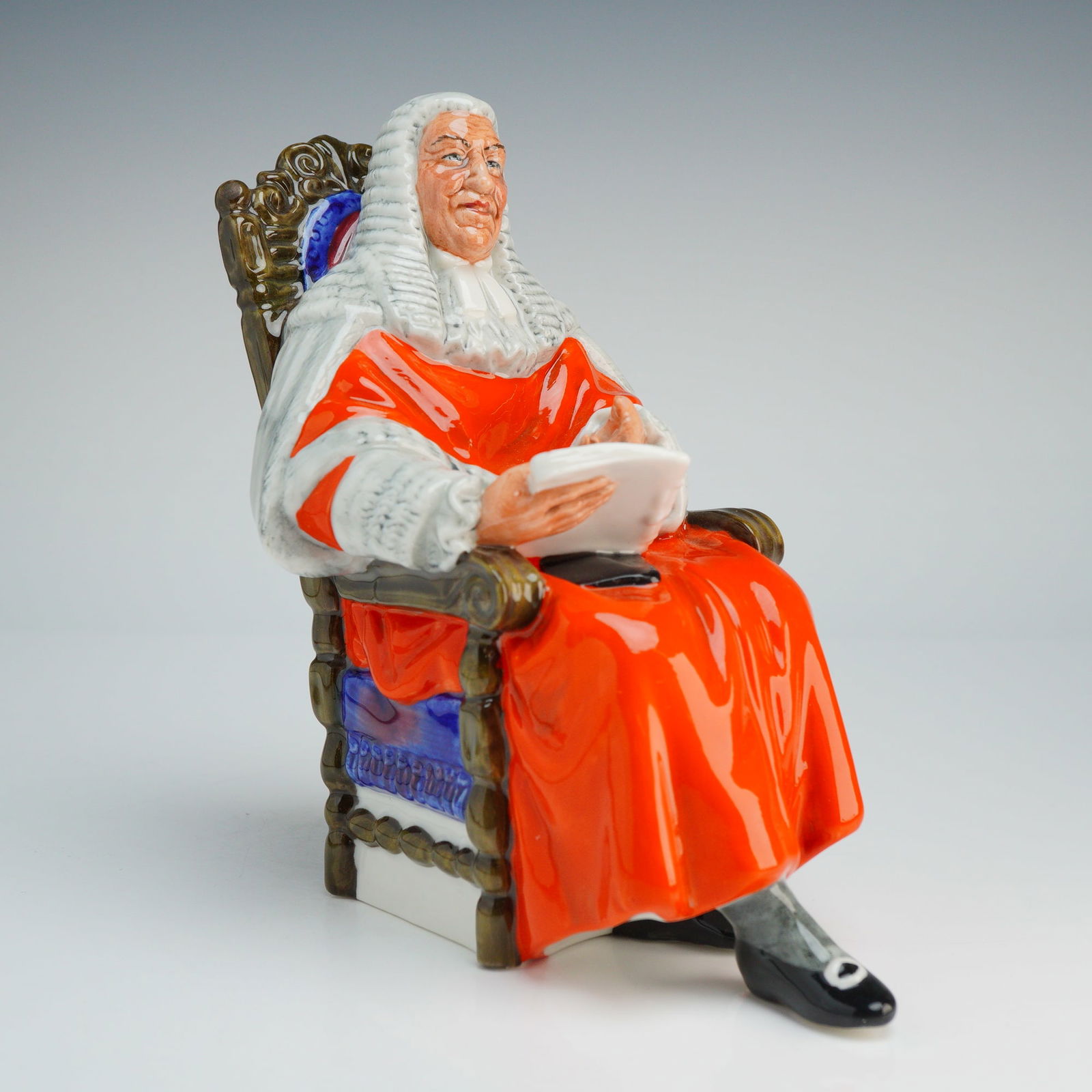 Royal Doulton Figurine, Judge HN2443: The figure depicts a seated male judge in traditional British legal attire, including vibrant red robes with white ermine-style trim, a full-bottomed white wig, and black buckled shoes. He is shown ho