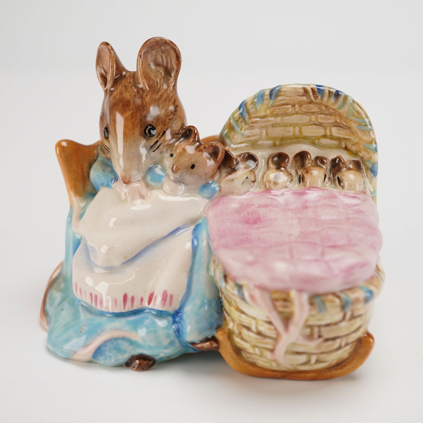 Beswick Beatrix Potter's Figurine, Hunca Munca: Designed by Arthur Gredington. The figure shows Hunca Munca seated in light blue and white clothing, holding an infant mouse beside a tan and brown wicker rocking cradle containing multiple infant mic