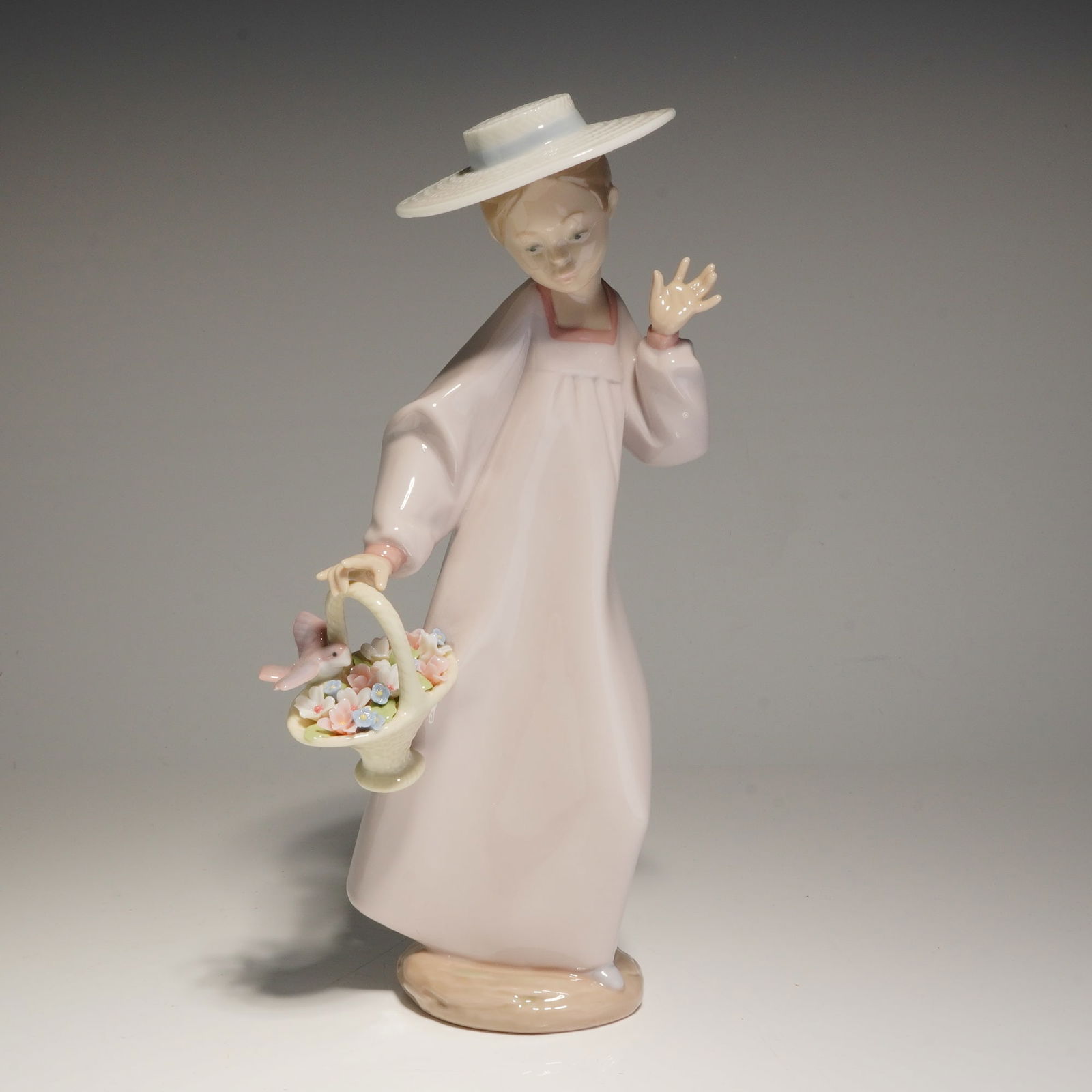 Lladro Porcelain Figurine, Hello Friend! 01006414: This Lladro figurine depicts a young girl wearing a wide-brimmed white hat with a blue band and a pale pink gown. The figure is posed waving with her left hand and carrying a basket of flowers in her