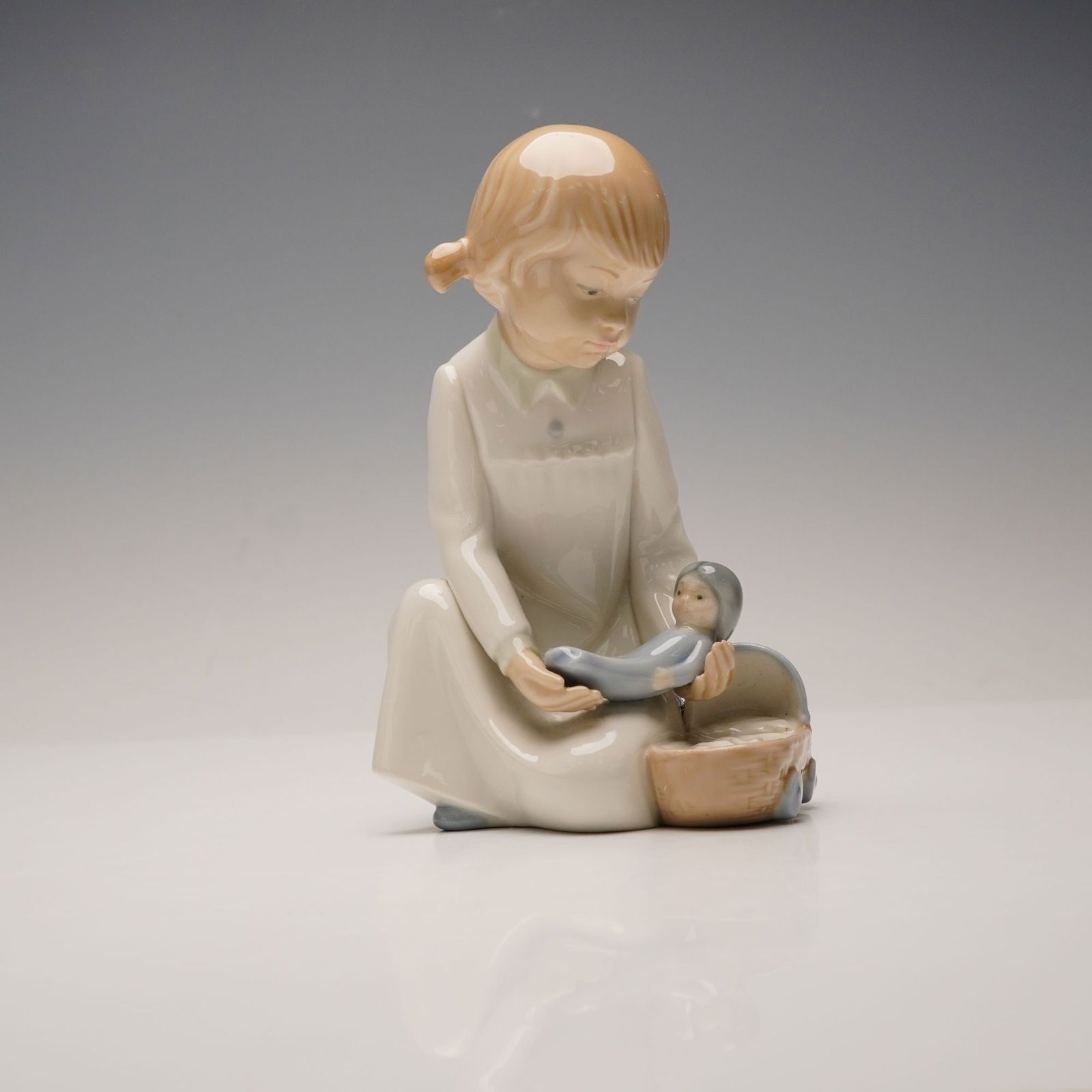 Nao by Lladro Porcelain Figurine, Girl with Rag Doll: This glazed porcelain figurine depicts a young girl with light brown hair in pigtails, wearing a cream-colored gown with pale blue slippers, seated and holding a small baby doll over a light brown and